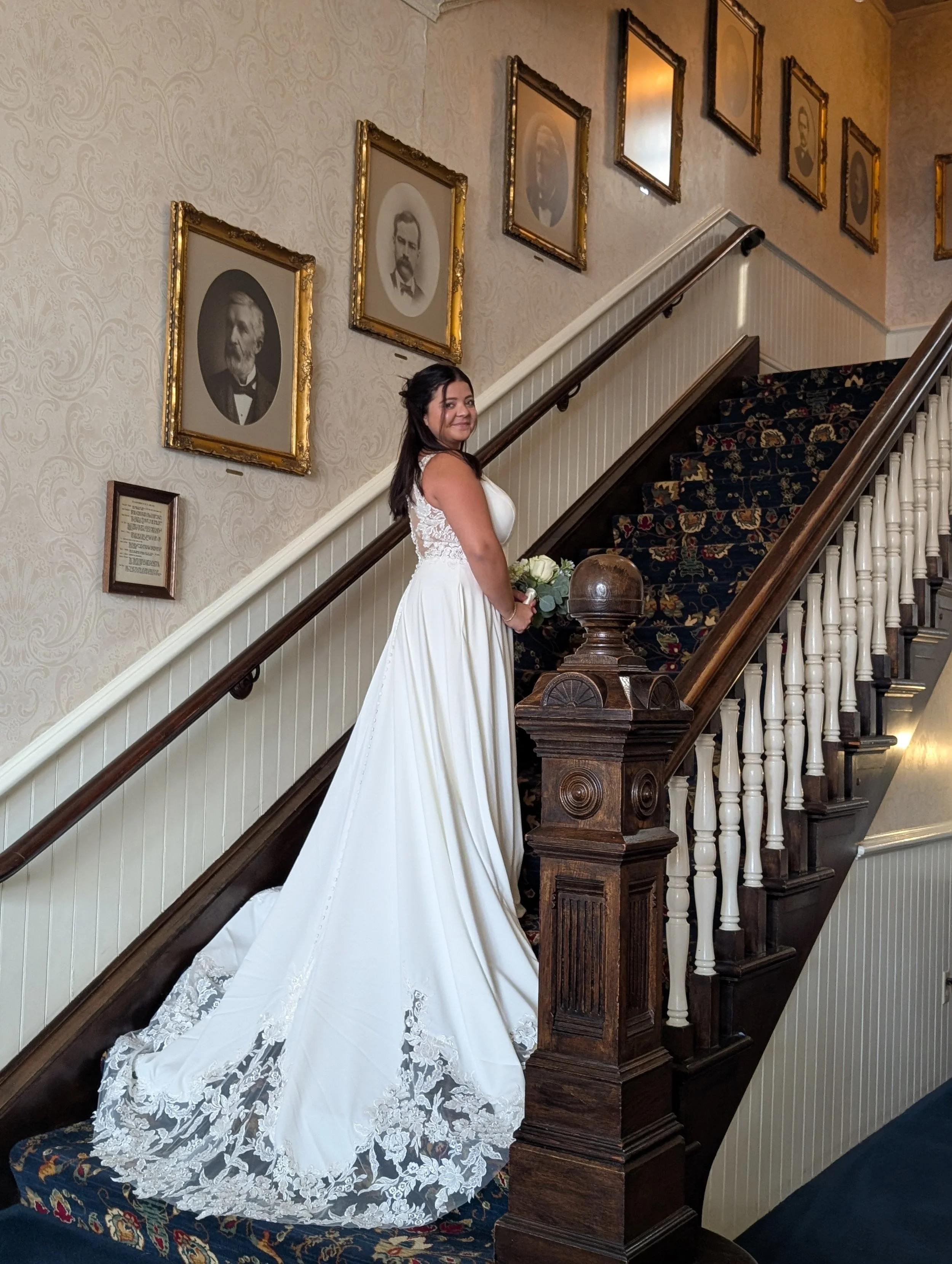 A bride in a white wedding dress standing on a staircase, holding a bouquet of white roses, indoors with framed black-and-white portraits on the wall behind her.