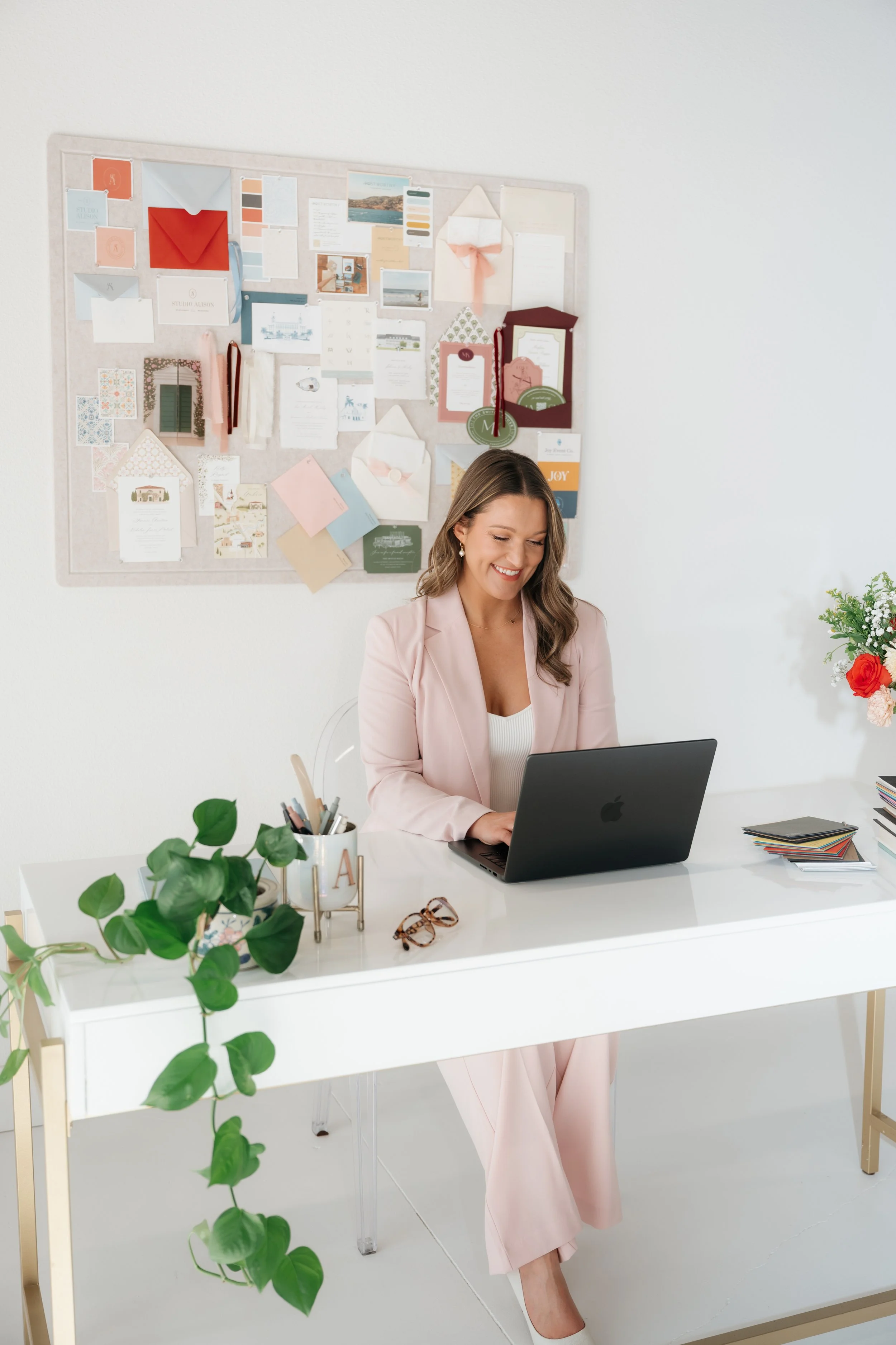 A woman sitting at a white desk, working on a laptop, smiling. The desk has a green potted plant, glasses, and stationery. Behind her is a bulletin board with various papers and decorations. The room has white walls and a bright, airy atmosphere.