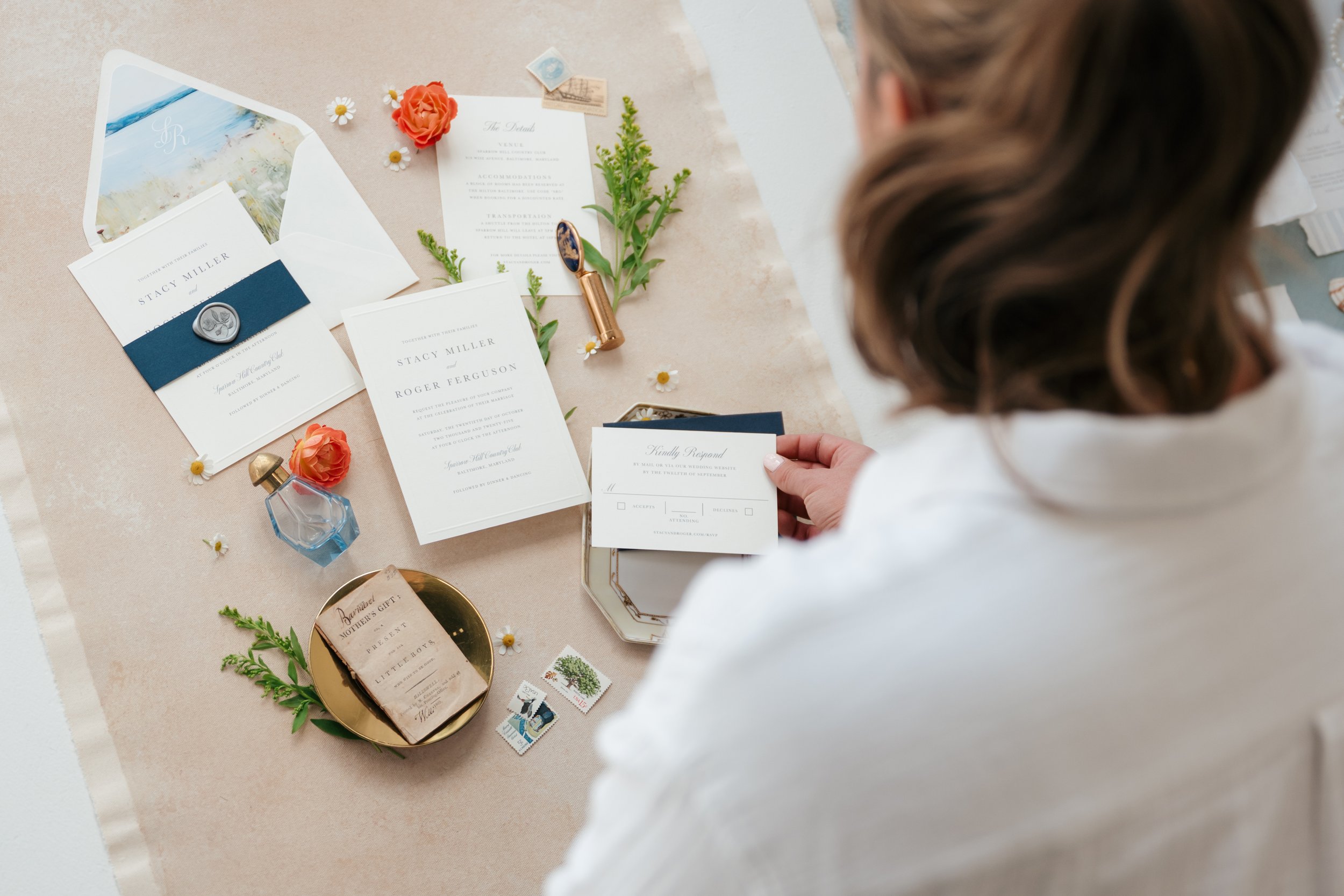 A person sitting at a table arranging wedding invitation materials, including invitations, envelopes, small decorative flowers, a perfume bottle, and stamps.