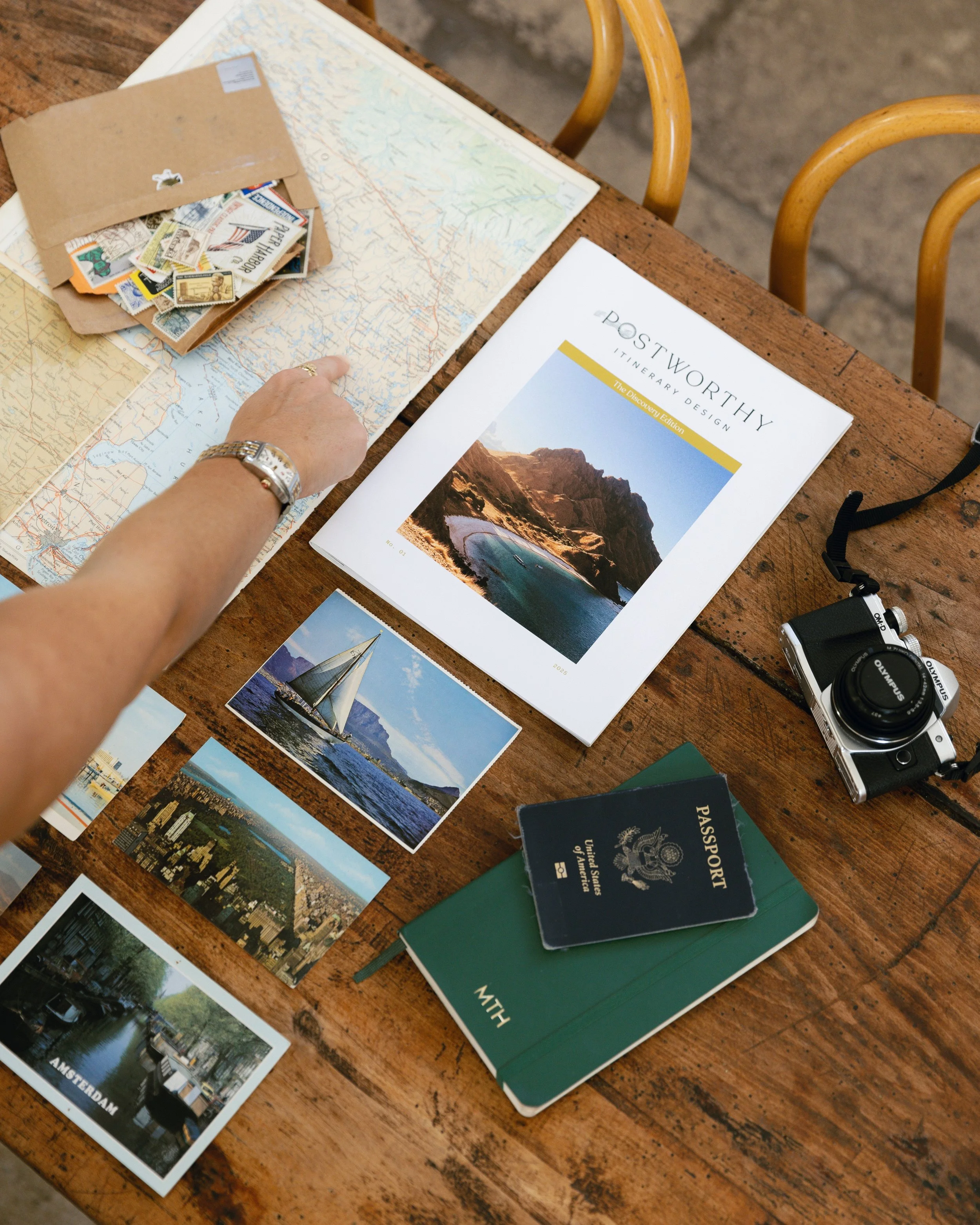 A wooden table with a travel planning setup including a map, a brochure titled 'Postworthy Itinerary Design,' photographs of a sailboat, cityscape, river, and a canal in Amsterdam, a passport, a green travel journal labeled 'With,' a camera, a folder of stamps, and a hand pointing at the map.