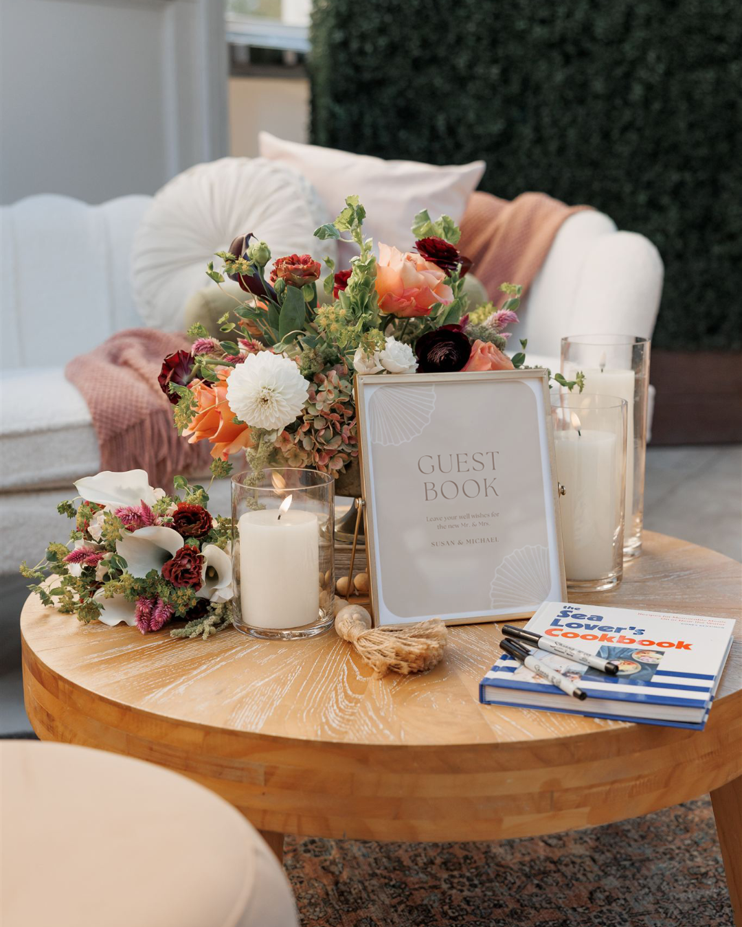 A welcome guest book and floral arrangement on a wooden table, with candles, a book, and markers in a cozy outdoor setting.