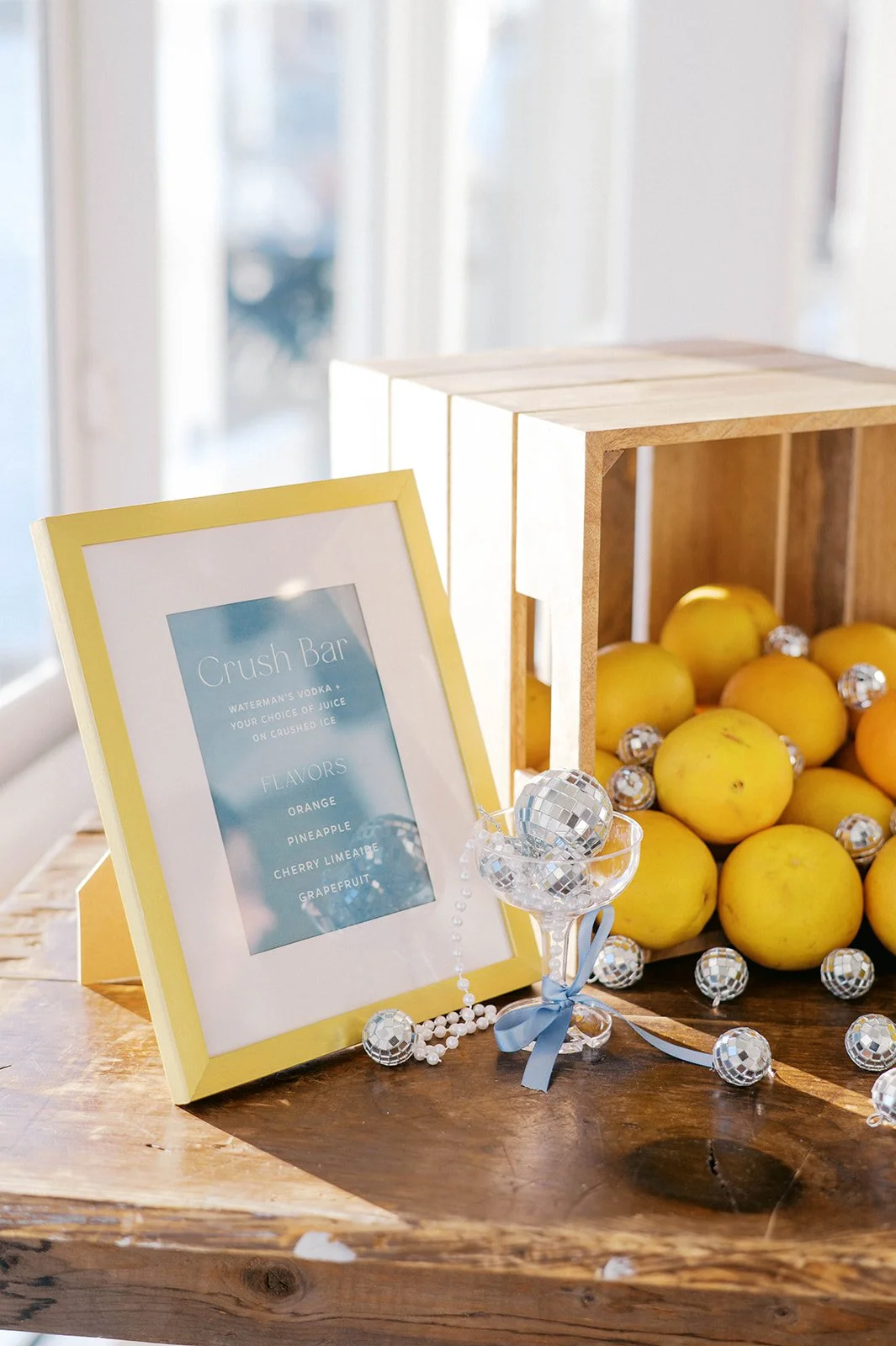 A table display featuring a framed sign for the 'Crush Bar' with a list of flavors including orange, pineapple, cherry limeade, and grapefruit, next to a glass bowl with a silver disco ball, surrounded by yellow apples and small silver disco balls, all placed on a wooden surface.