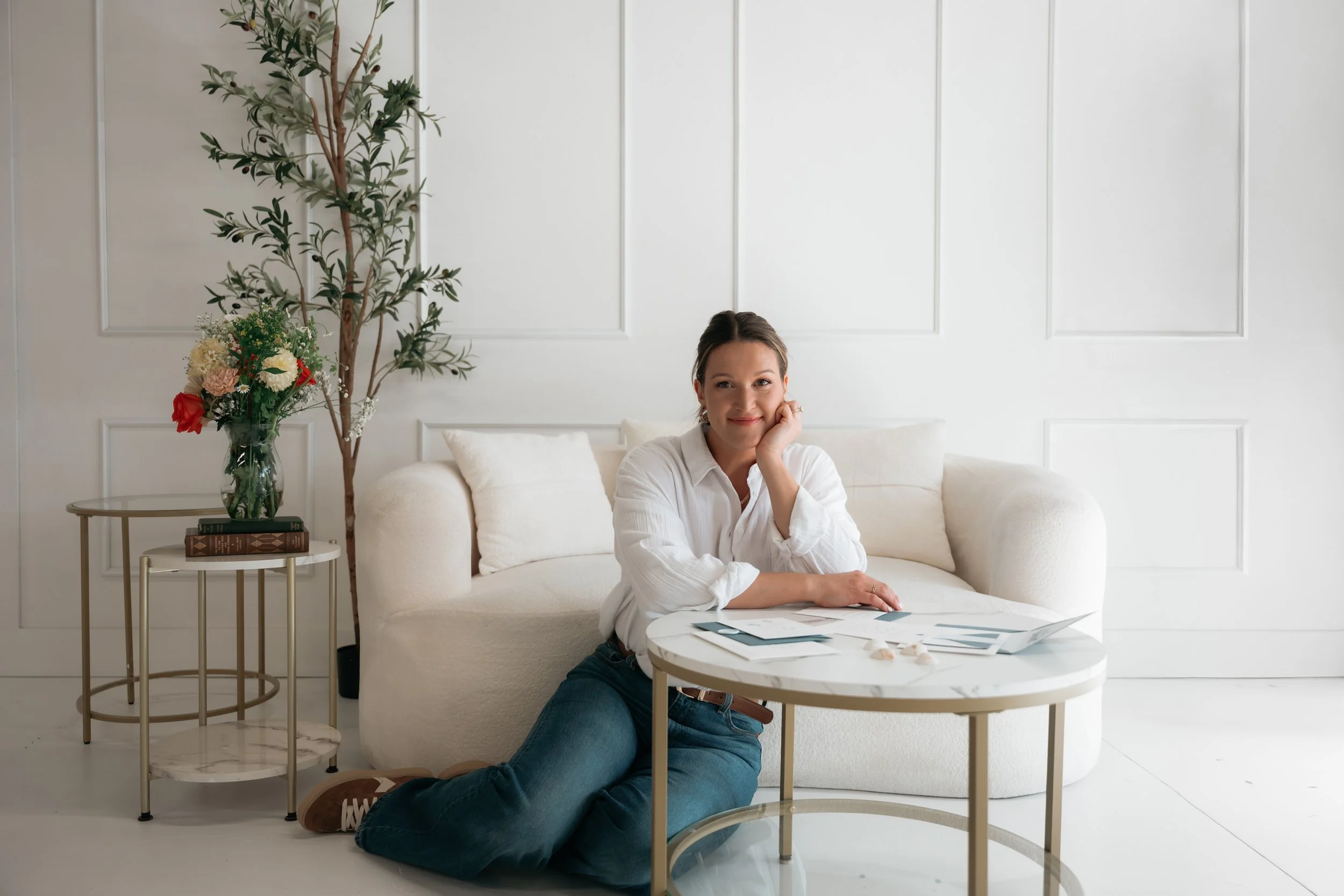 A woman sitting on the floor leaning against a white sofa with her arm resting on a round coffee table, smiling. There is a side table with a vase of flowers and books, and a tall plant in the background.