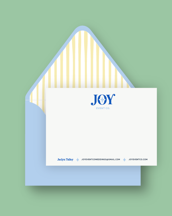 A white business card with the word 'JOY' in blue and a star inside the letter 'O', placed in front of a blue envelope with yellow and white striped lining, on a green background.