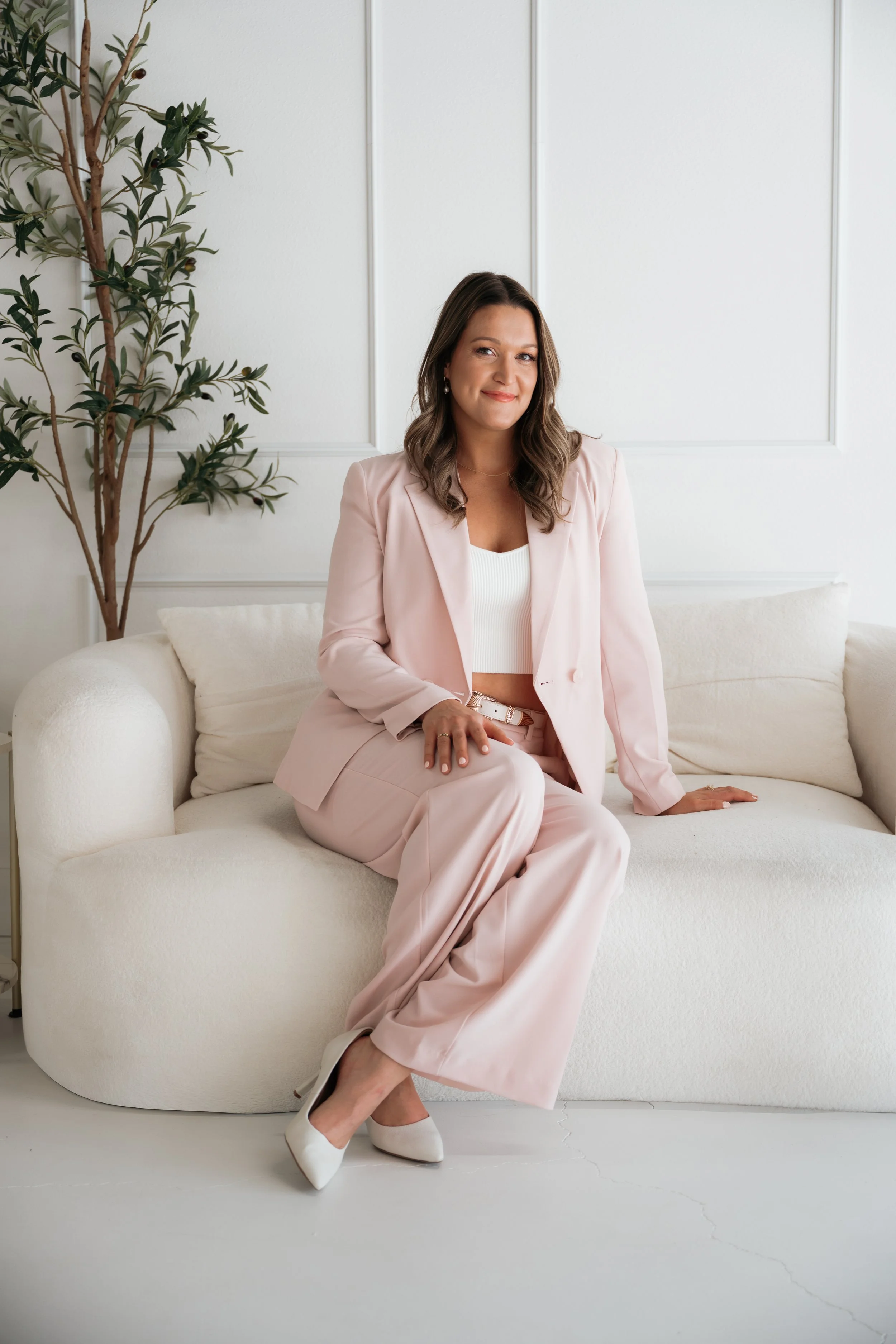 A woman sitting on a white couch in a bright, minimalist room. She is wearing a light pink suit and white high heels, smiling at the camera with a potted plant behind her.