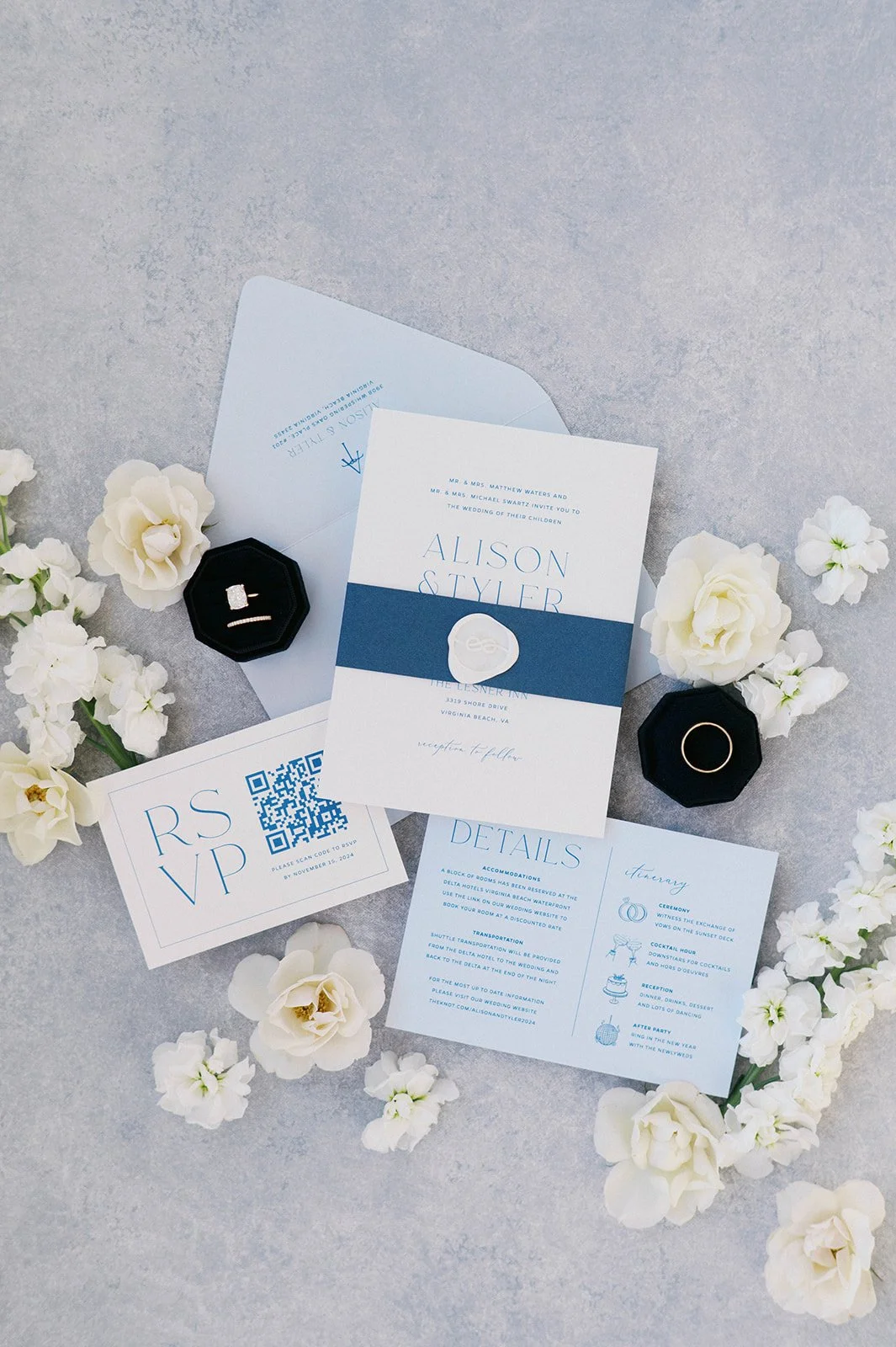 Wedding invitation set with a blue and white color scheme, surrounded by white flowers and black ring boxes containing rings, including a diamond engagement ring and wedding band.