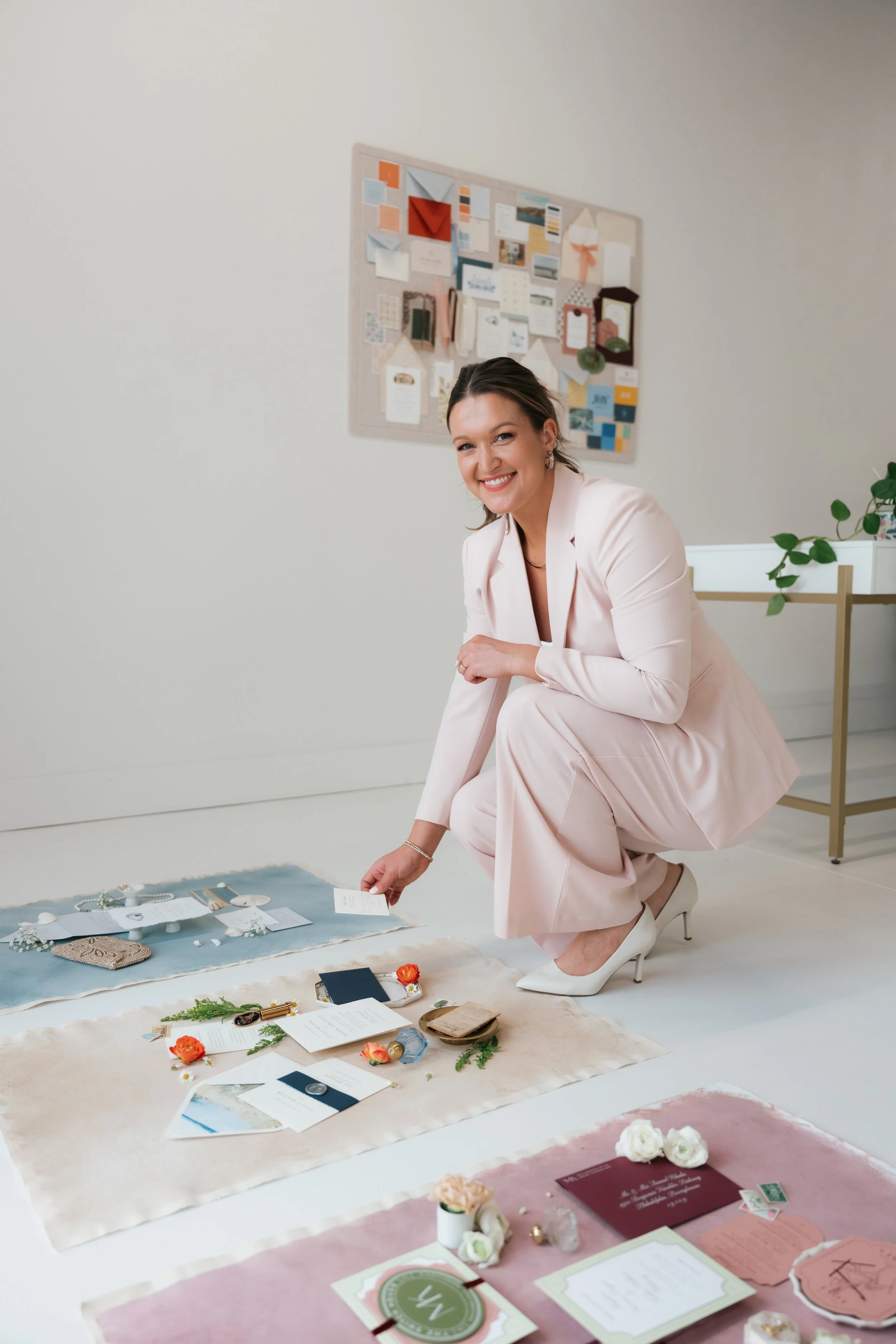 Woman in a pink suit kneeling on the floor, smiling, surrounded by wedding invitations, flowers, and decorative items for a wedding or event.
