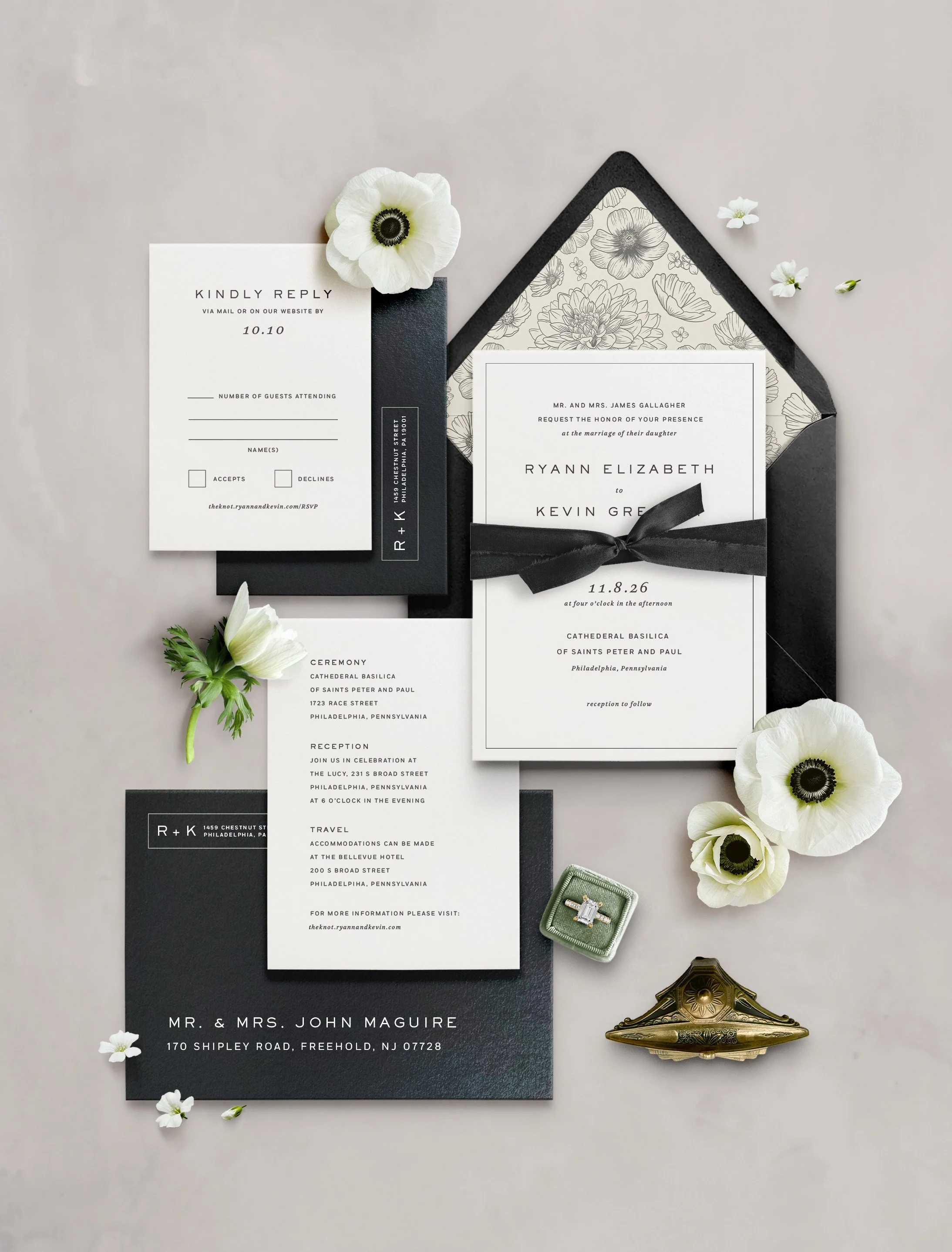 Wedding invitation suite including an invitation card, RSVP card, details card, all in black and white with floral accents, accompanied by flowers, a ring box, and a decorative item.