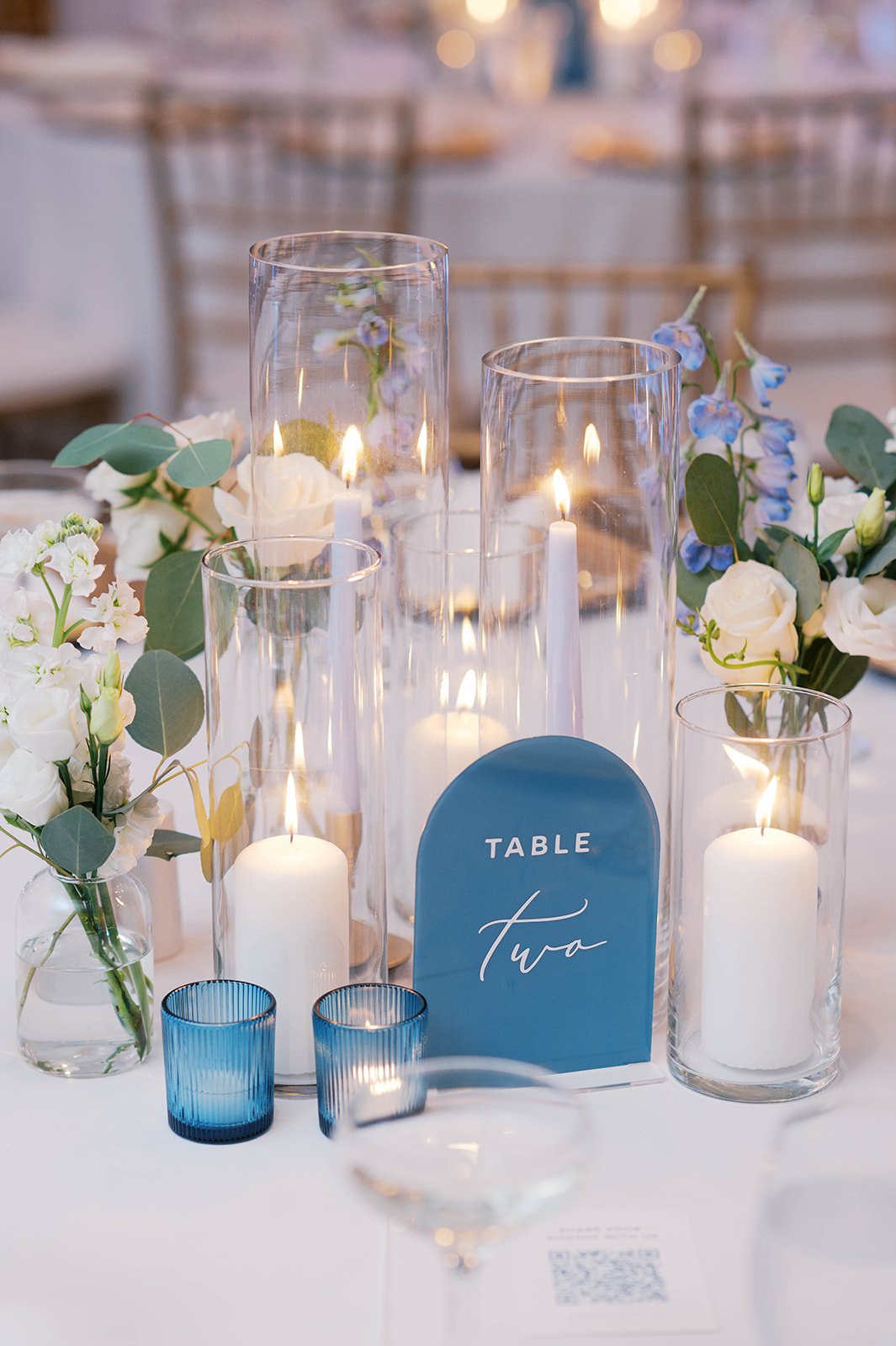 Wedding table centerpiece with white and purple flowers, green foliage, candles in glass holders, and a blue table sign that says 'Table Two'.
