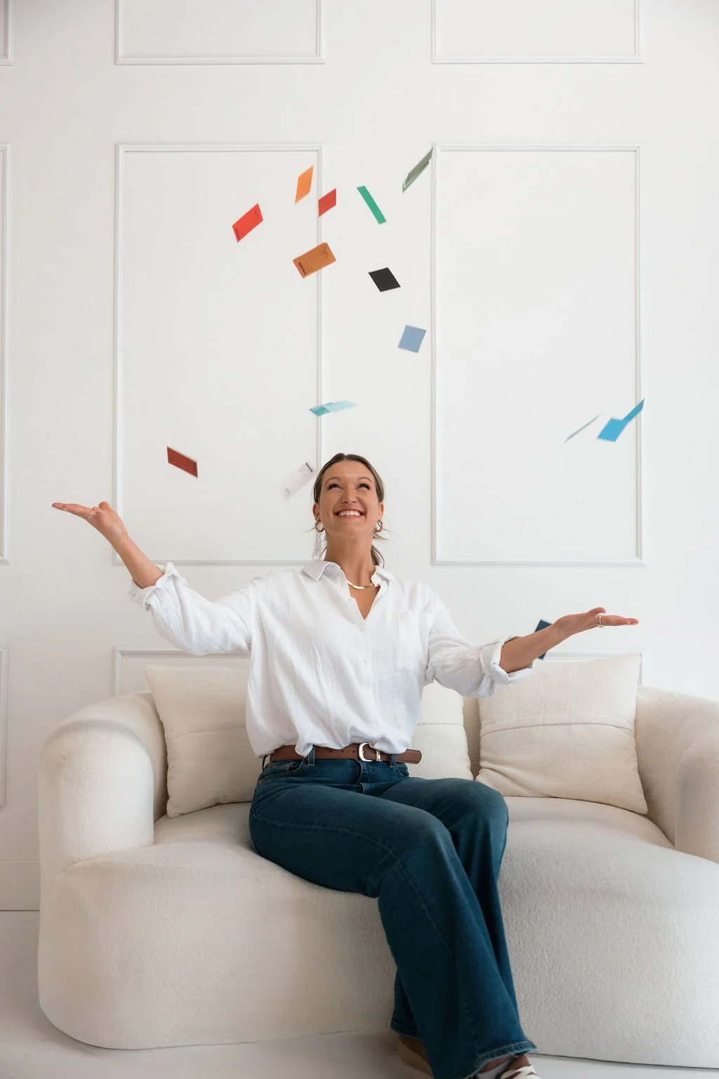 Woman sitting on a white couch, smiling and throwing colorful paper notes in the air indoors.