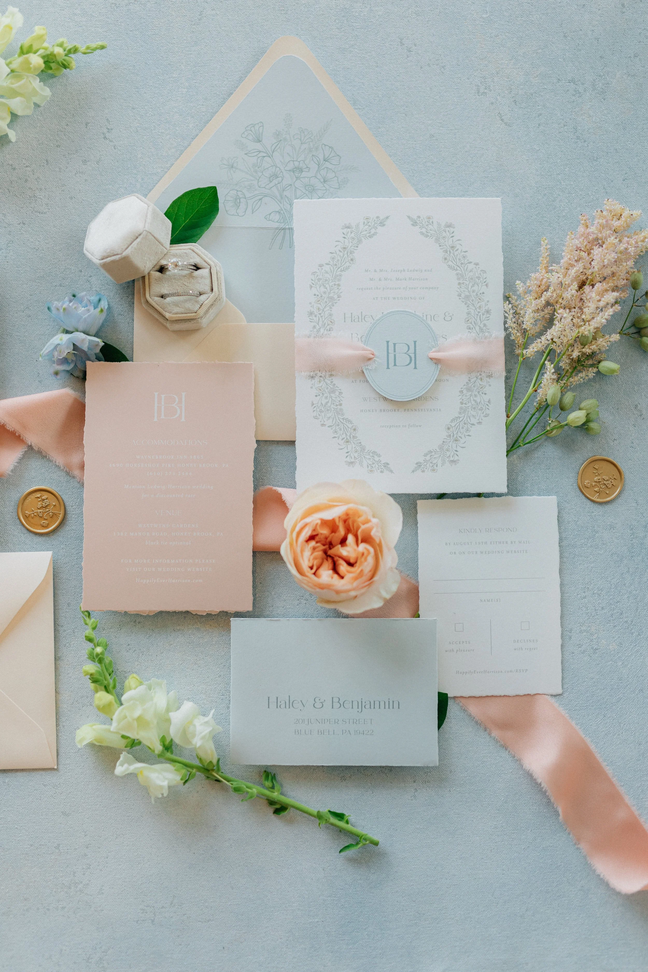 Flat lay of wedding invitation suite with floral decorations, ribbons, and wedding rings in a small box on a light fabric background.