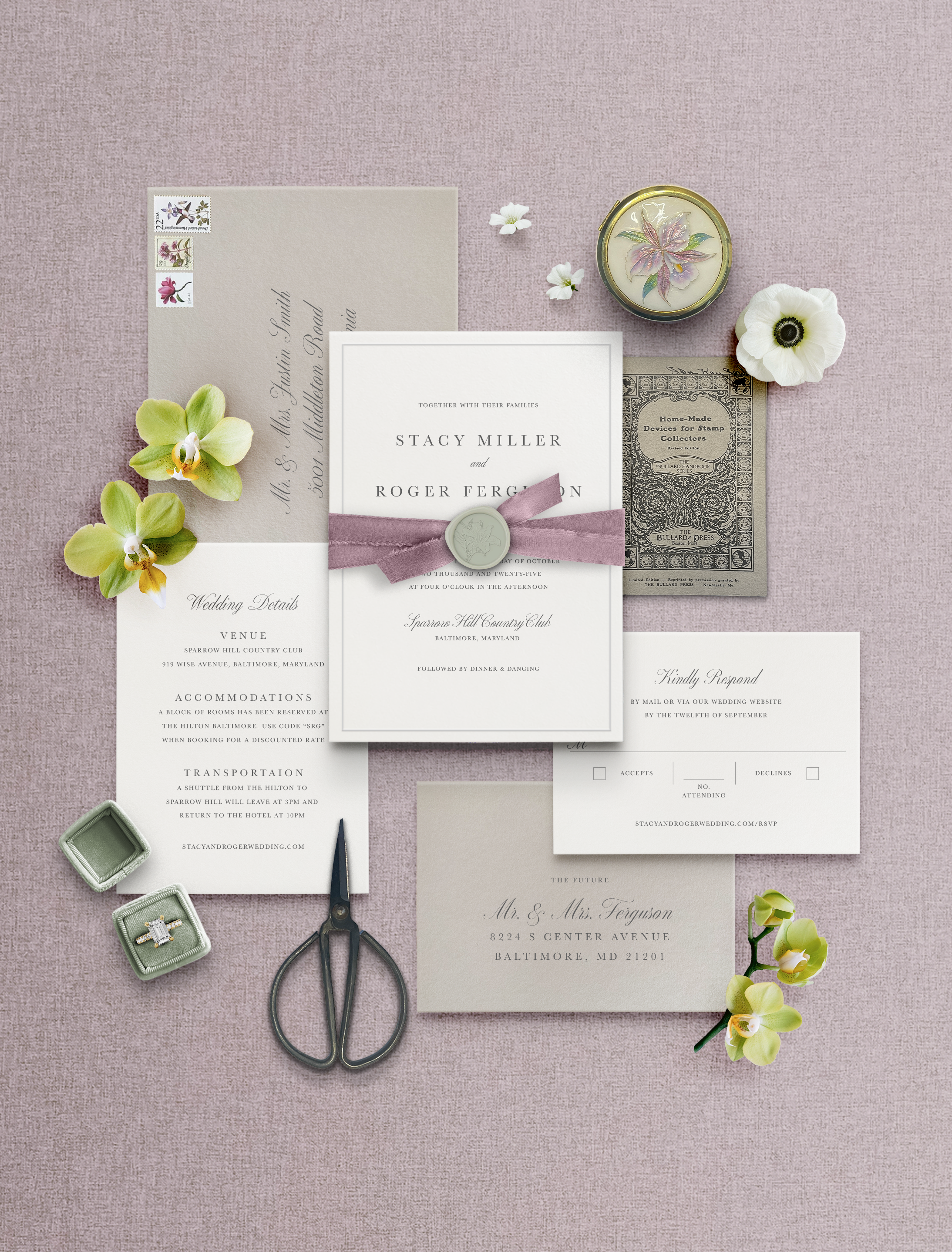 Wedding invitation suite with floral accents, stamps, a pair of scissors, rings in a box, and decorative items, all arranged on a pink textured surface.