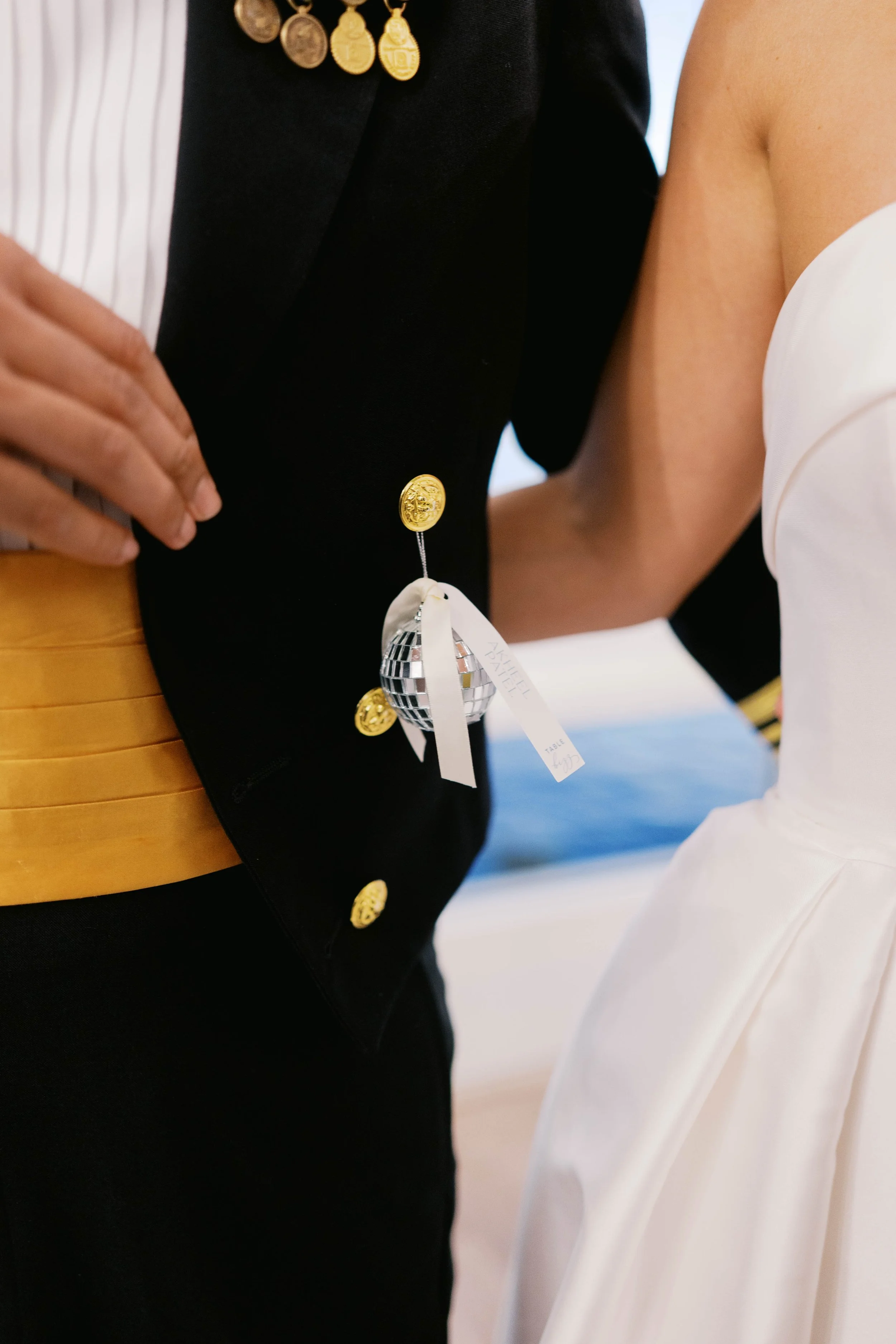 Close-up of two people at a formal event, one wearing a black jacket with gold buttons and medals, and the other in a white dress. The person in the black jacket has a decorative pin shaped like a disco ball with a white ribbon attached.