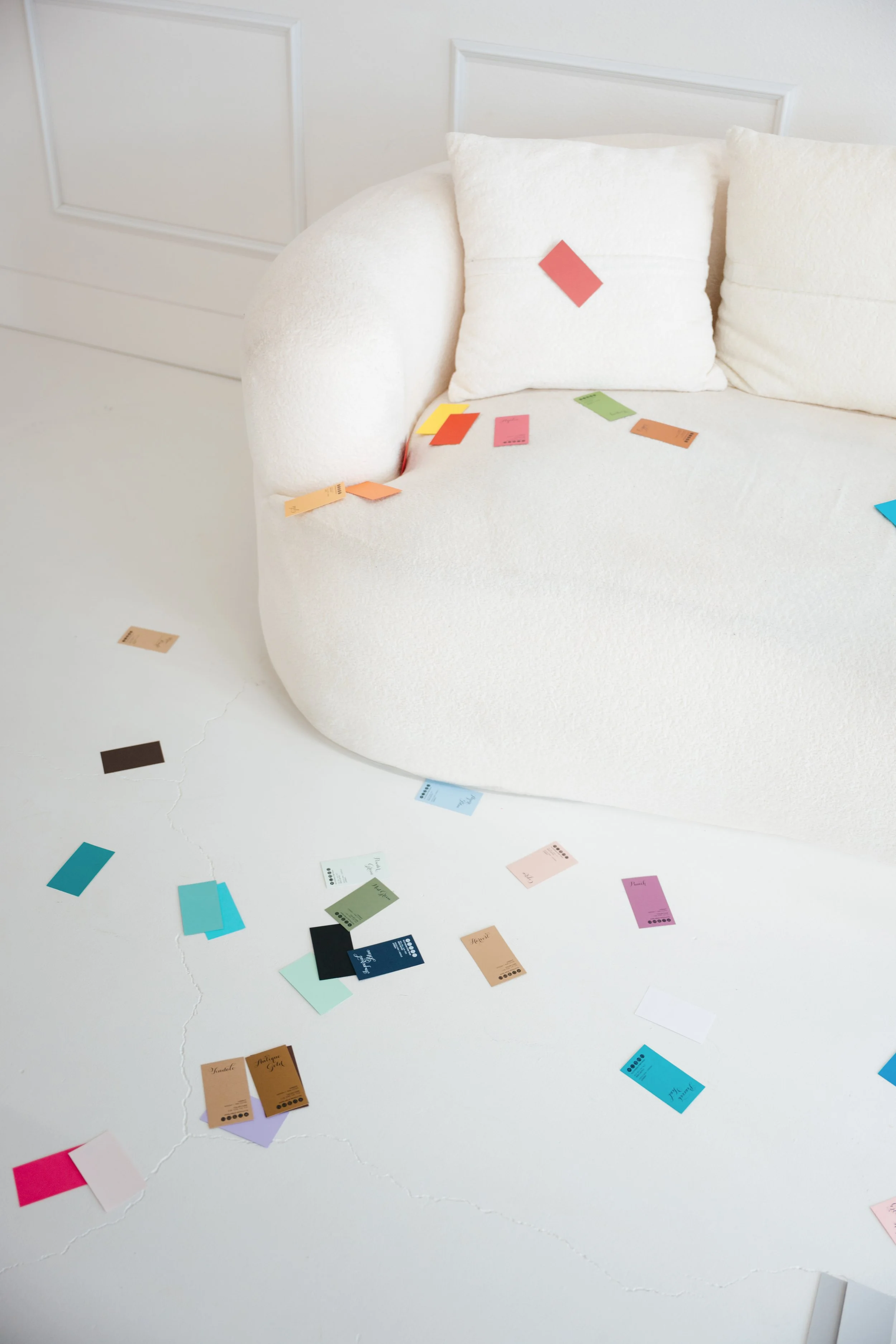 A white, curved sofa with a white pillow, with colorful paper swatches scattered on the sofa and floor. The background features a white wall with paneling.