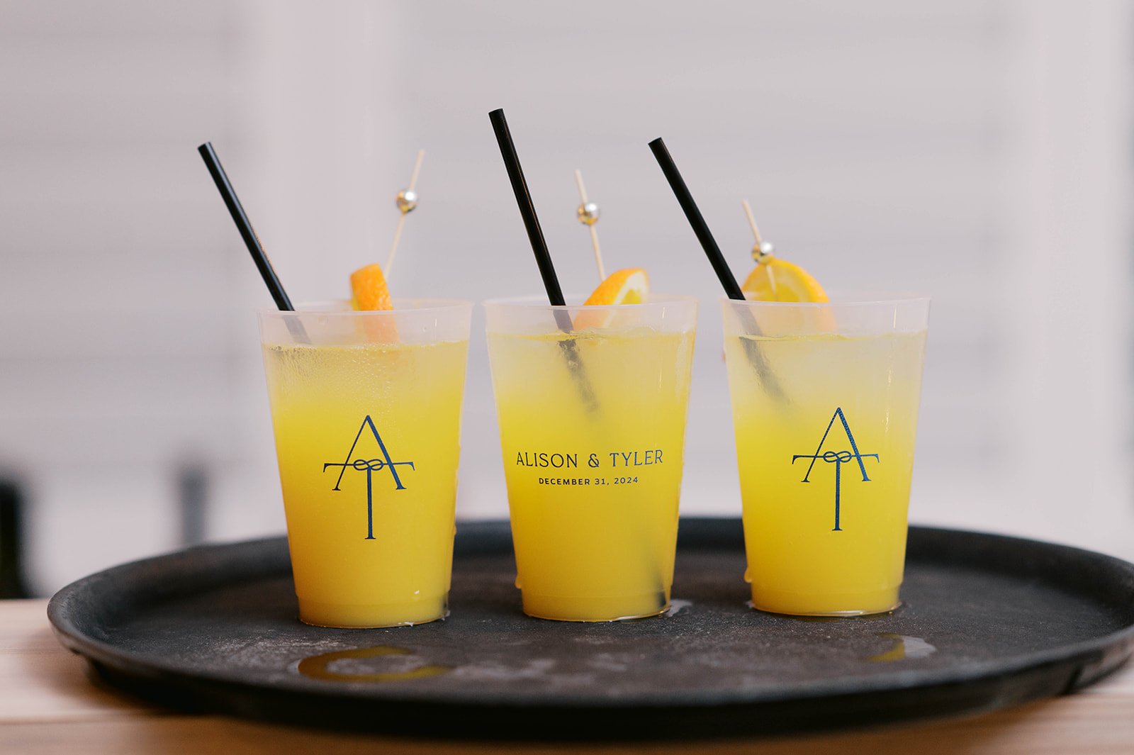 Three tall glasses of yellow lemonade with lemon slices, black straws, and decorative picks, placed on a black tray.