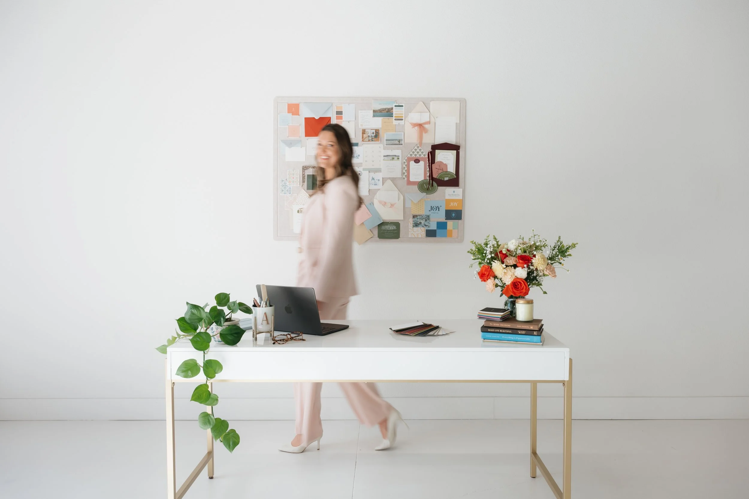 A woman in a pink suit walking past a white desk with a laptop, potted plant, stack of books with a vase of flowers on top, and a color swatch book. There is a bulletin board with cards and decorations on the white wall behind her.