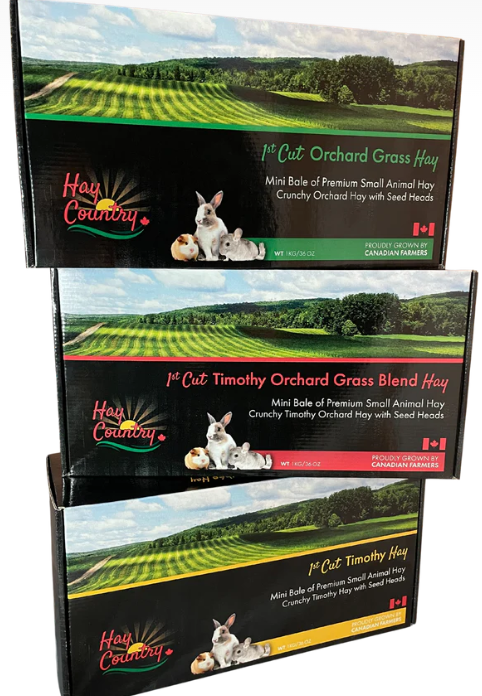 Three boxes of orchard grass hay stacked, labeled as 1st Cut Orchard Grass Hay, 1st Cut Timothy Orchard Grass Blend Hay, and 1st Cut Timothy Hay, each with images of farm landscapes and small animals like rabbit and guinea pig on the packaging.