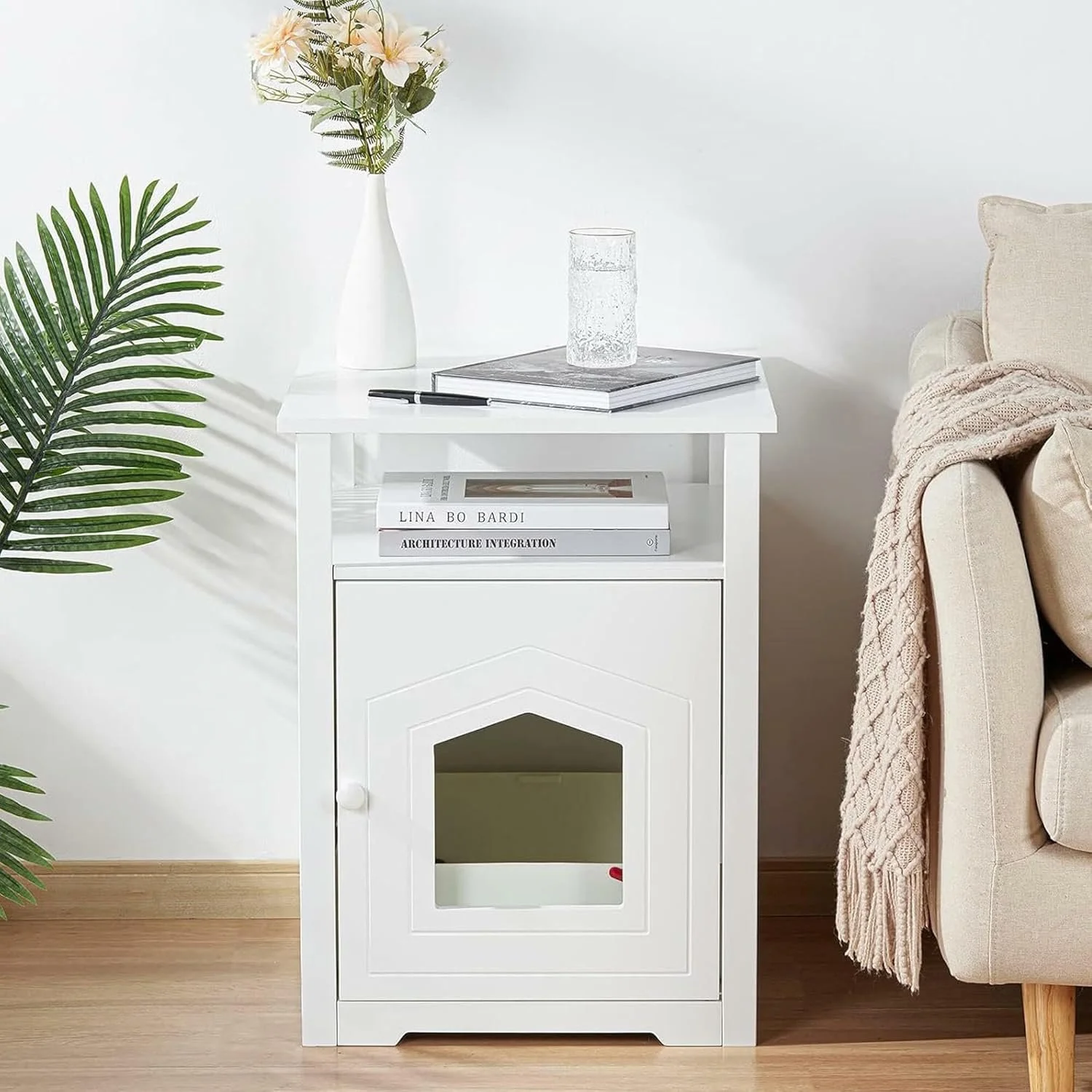White side table with books, a glass of water, and a vase with flowers next to a beige sofa with a throw blanket. A green plant is partially visible on the left.
