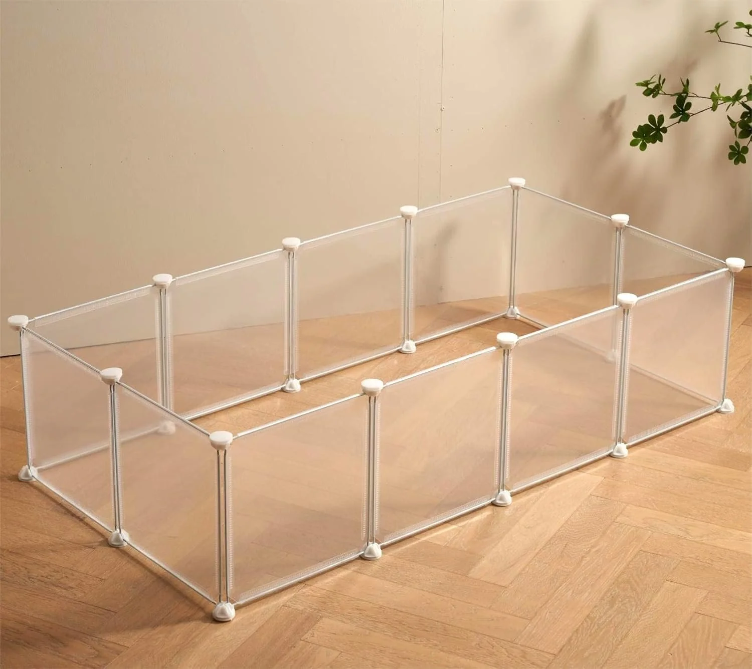 A grey, multi-compartment plastic storage organizer with transparent panels and white connectors, placed on a wooden floor near a white wall and a green plant.