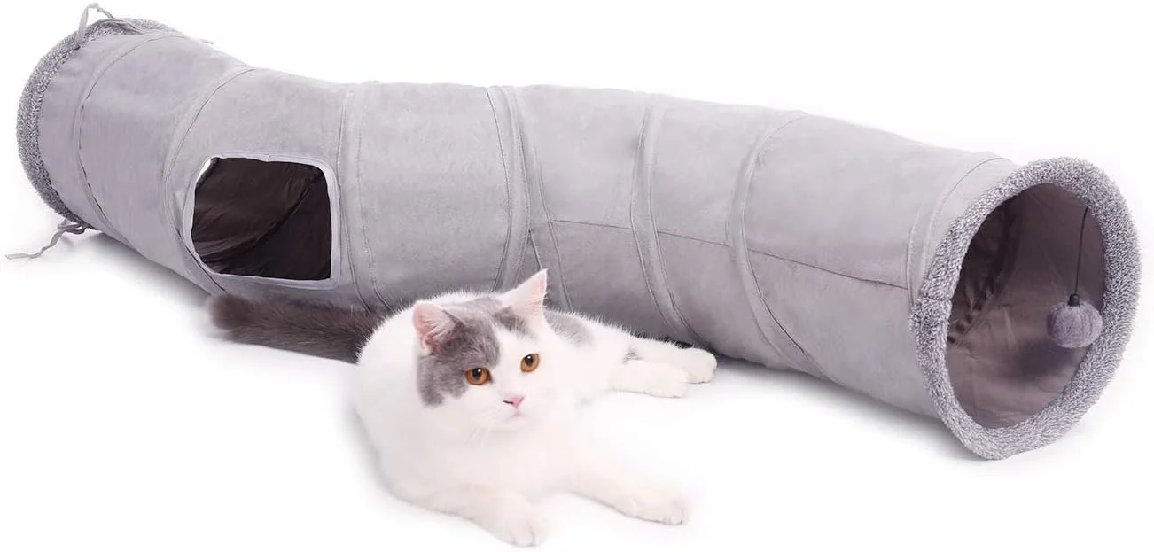 A white and gray cat lying next to a gray fabric cat tunnel with a small entrance and a dangling pom-pom toy inside.