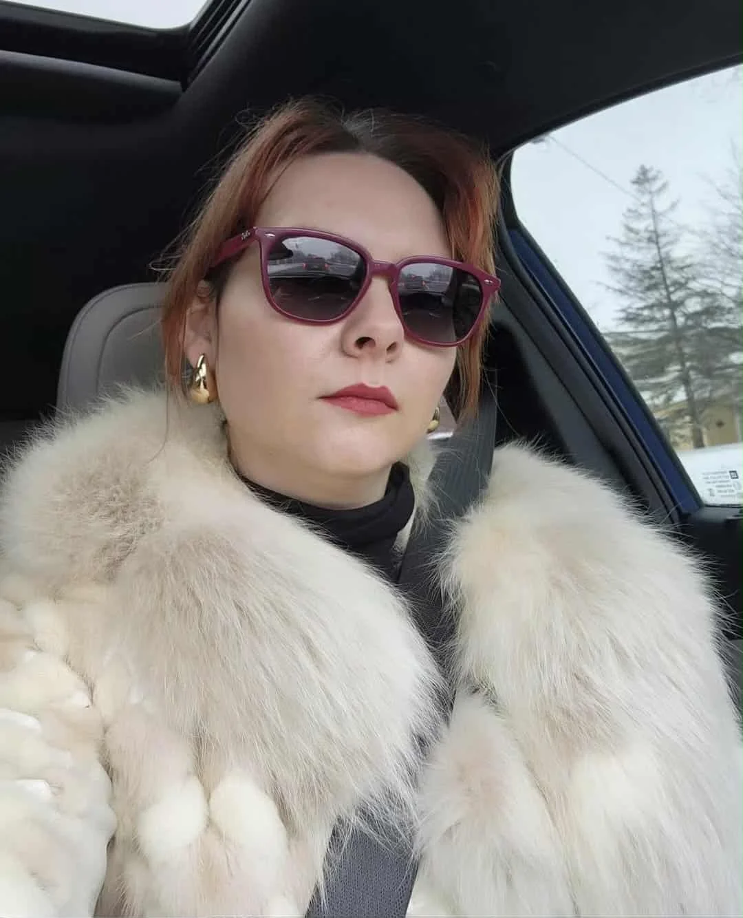 Woman with reddish hair wearing sunglasses, gold earrings, and a fur coat, sitting in a car with a snowy landscape visible through the window.