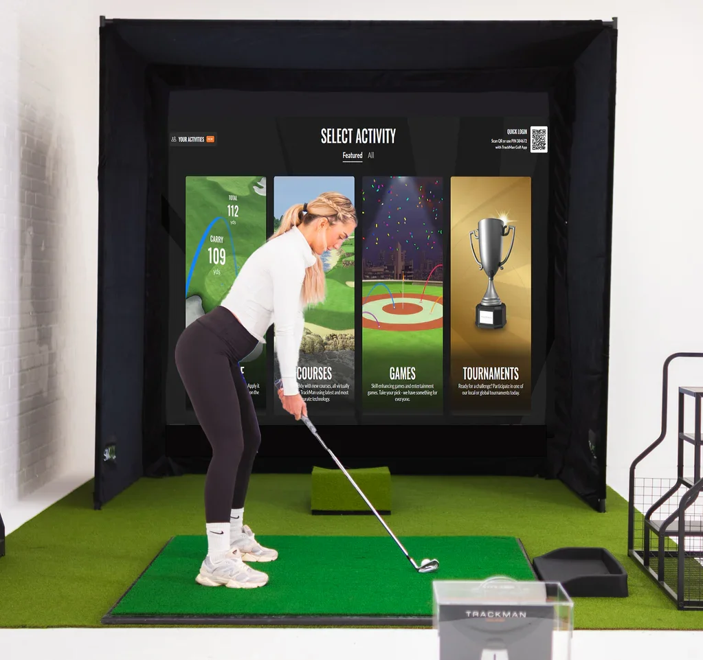 A woman playing indoor golf on a simulation screen, dressed in golf attire, using a golf club on a green mat.