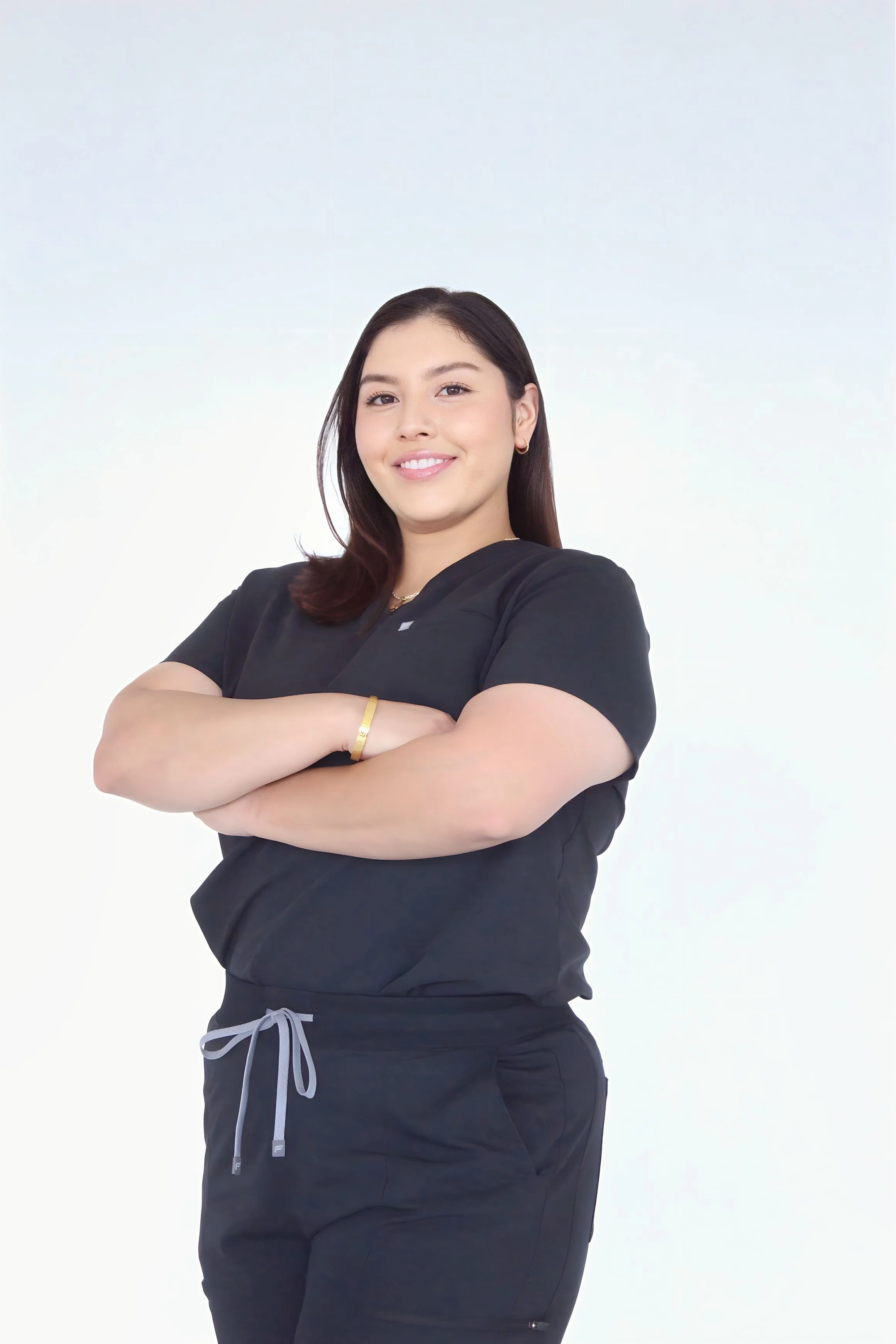 A young woman in black medical scrubs standing with arms crossed, smiling confidently against a white background.