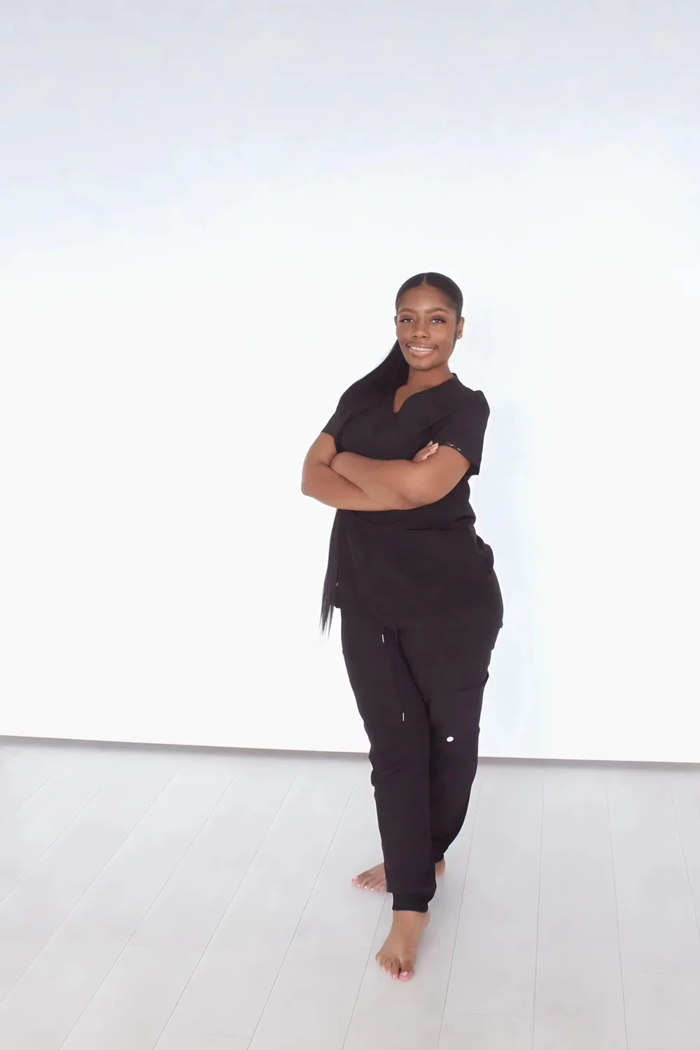 A woman in black scrubs standing with arms crossed, smiling in front of a plain white wall.