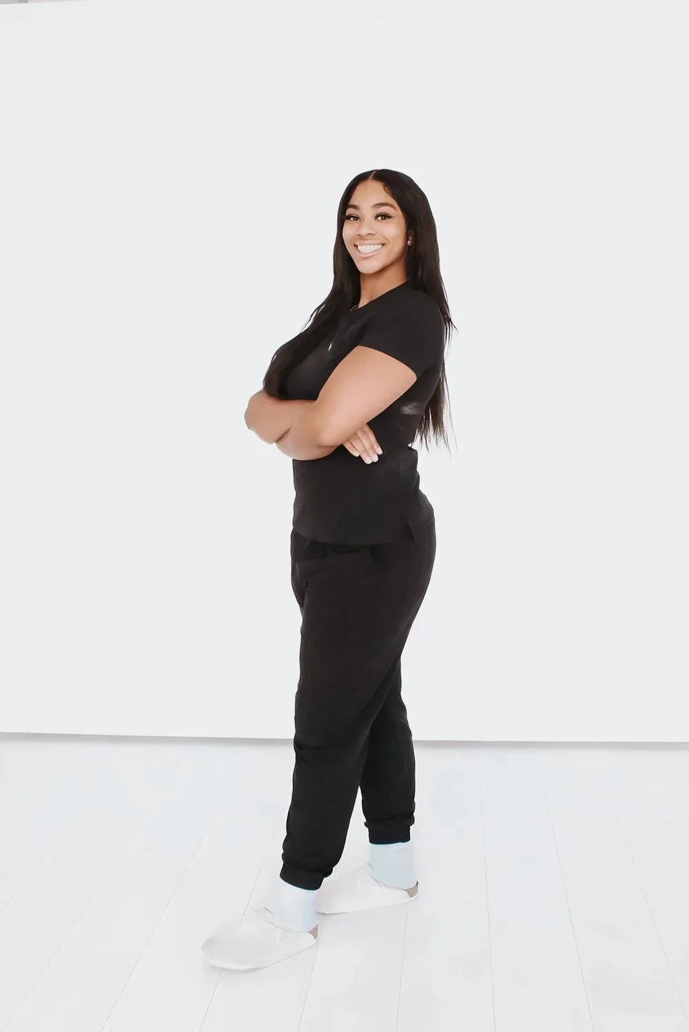 Young woman with long dark hair wearing black casual clothes, standing with arms crossed and smiling, against a plain white background.