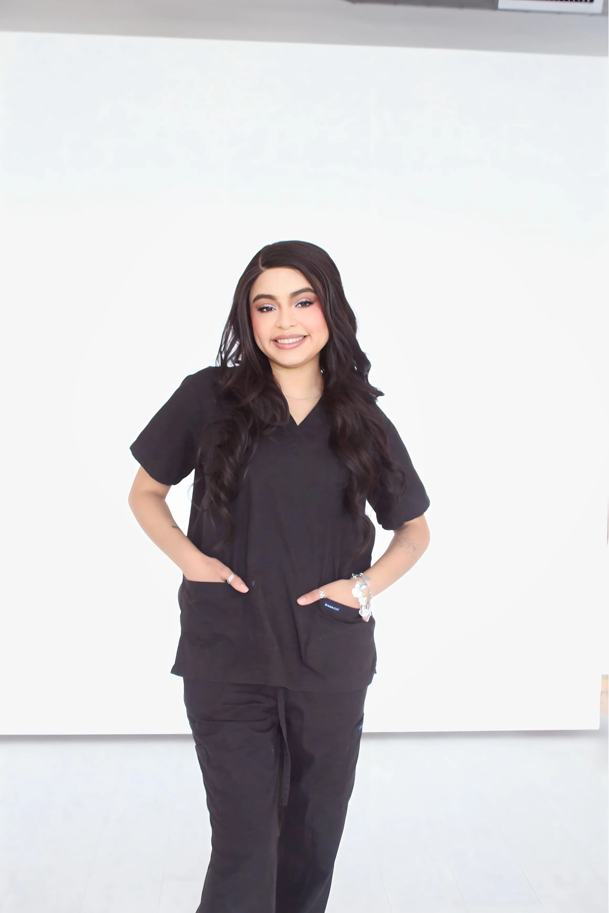 A woman with red hair tied up, wearing black scrubs, standing against a white background with a friendly smile.