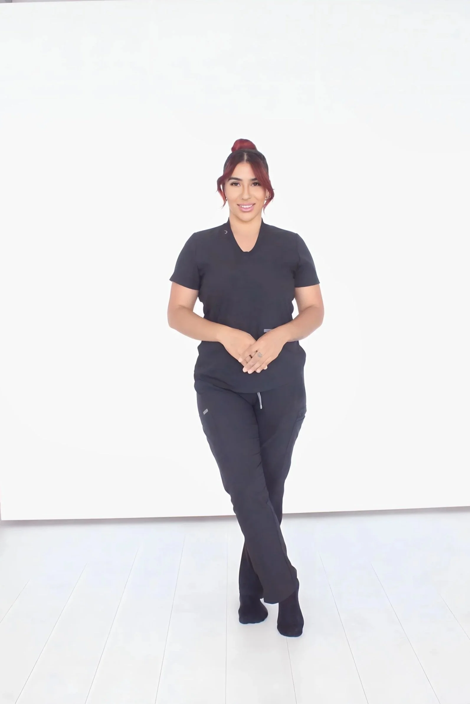 A woman with red hair tied up, wearing black scrubs, standing against a white background with a friendly smile.