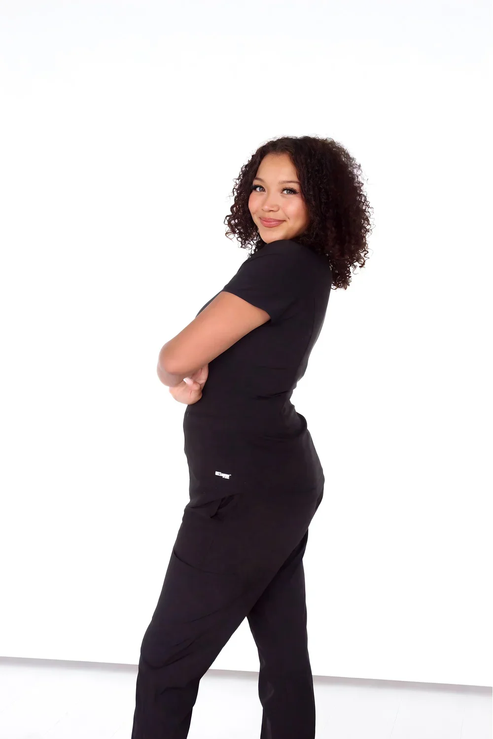 Young woman with curly hair wearing black scrubs, standing with arms crossed, smiling, against a white background.