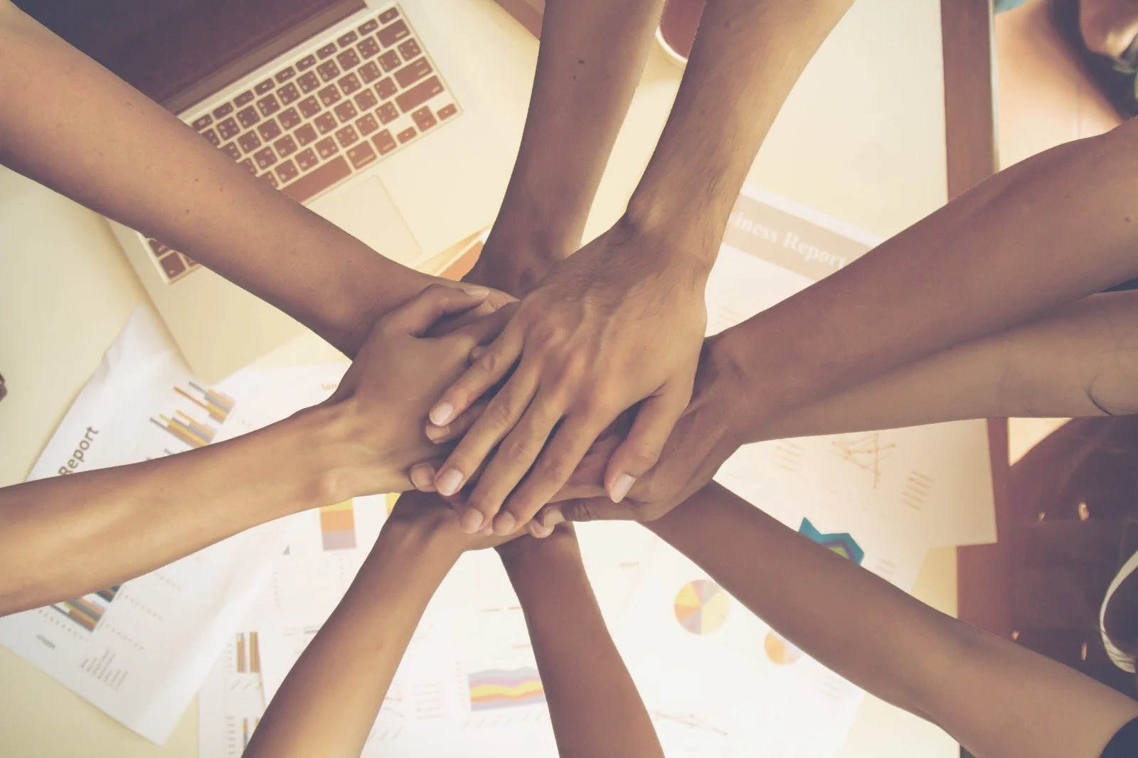 Multiple hands from diverse individuals stacked together in a show of teamwork over a desk with papers, a laptop, and business reports.