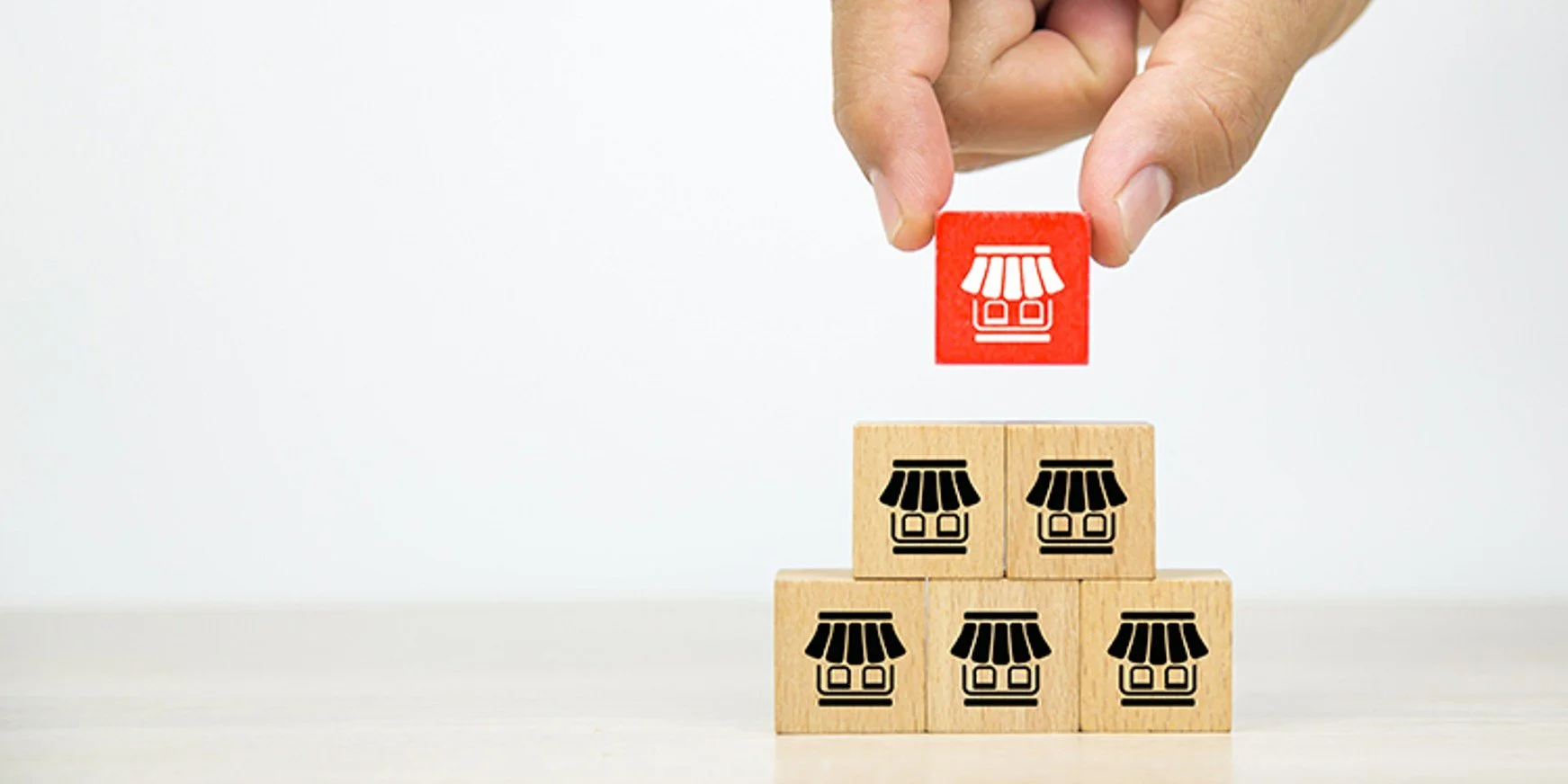 A person's hand placing a red block with a store icon onto a stack of five wooden blocks with matching store icons, arranged in a pyramid.