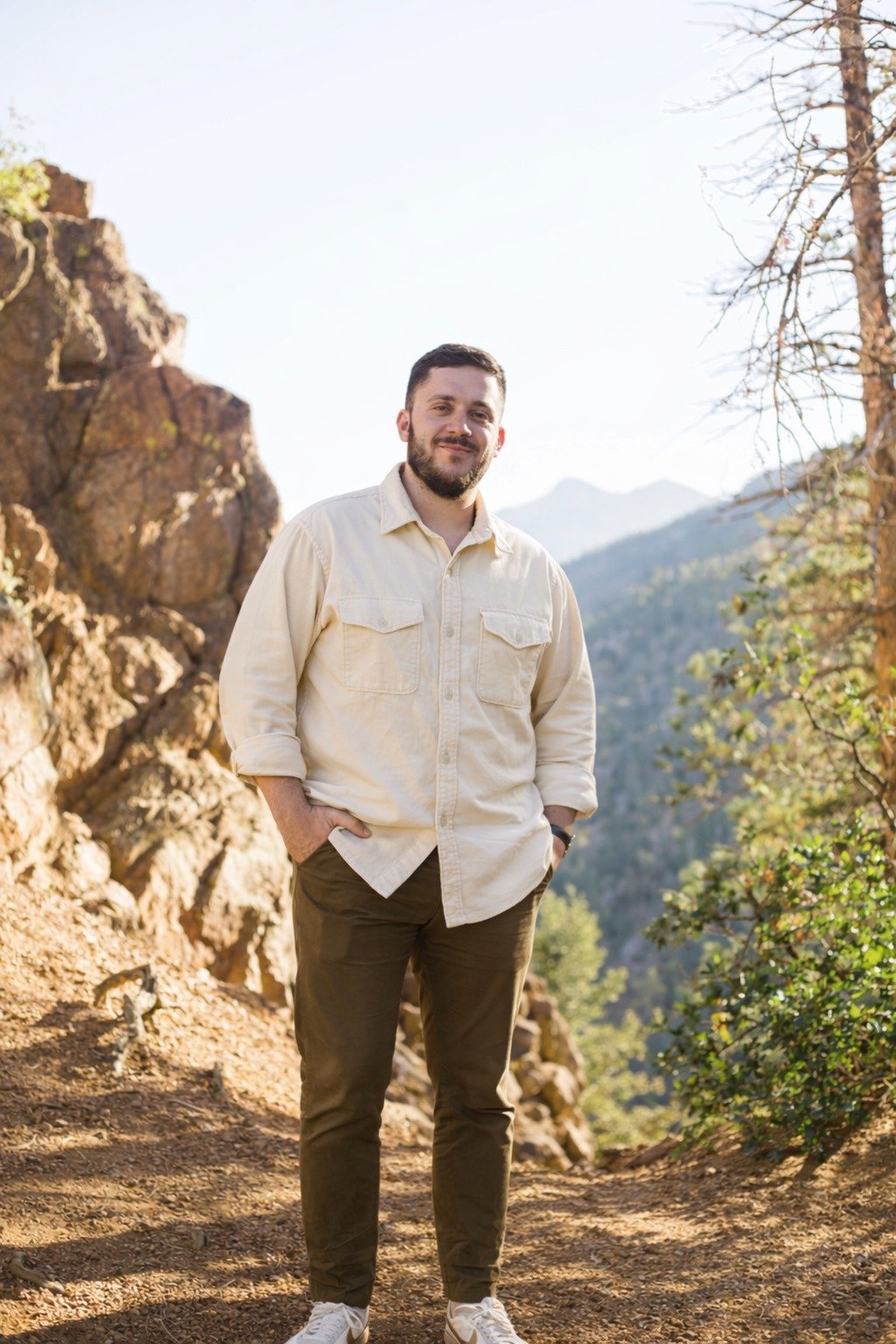 A man standing outdoors on a trail with rocky mountains and trees in the background, smiling and wearing a light-colored shirt and dark pants.