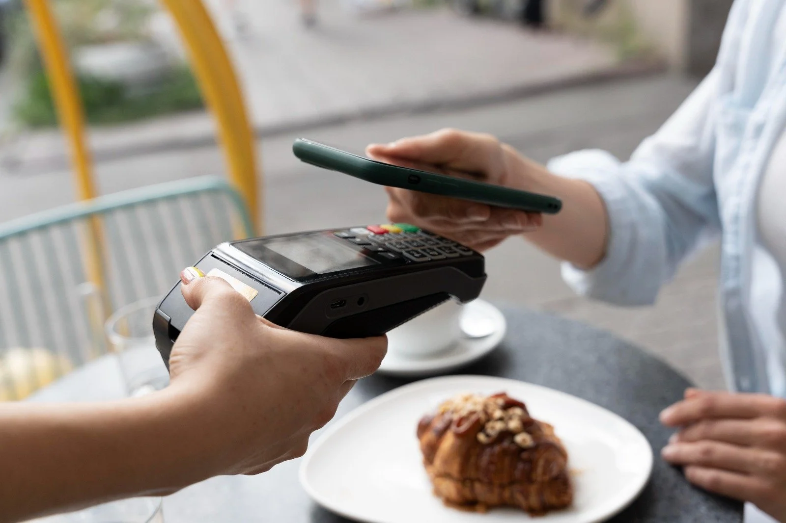 Person using a mobile phone to pay at a restaurant counter with a point-of-sale terminal, a pastry on a white plate, and a cup of coffee in the background.
