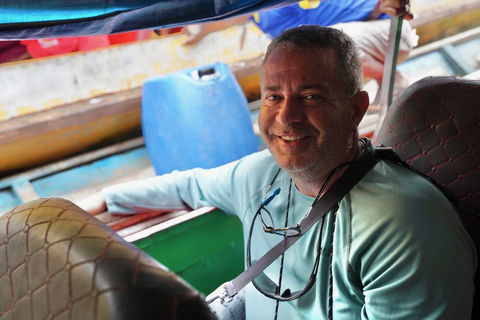 Smiling man with short gray hair, wearing a light blue shirt, sitting on a boat with colorful boat seats and large blue container in background.