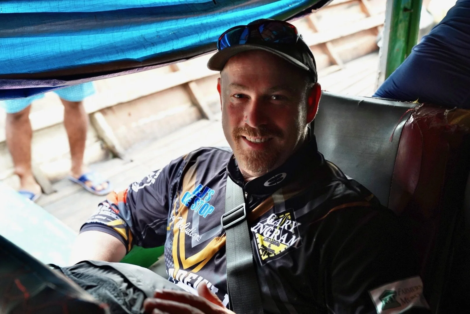 Man in a black and gold cycling jersey with patches, smiling and sitting inside a vehicle, with sunglasses on his forehead.