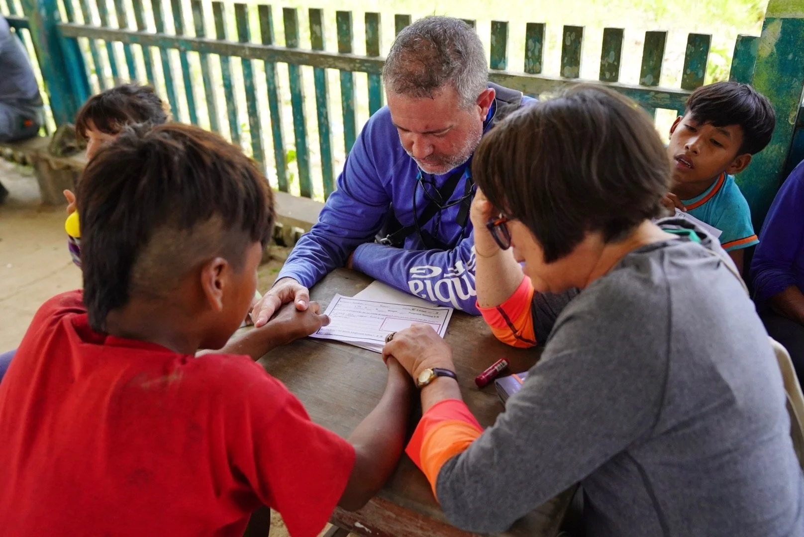 A diverse group of children and two adults sitting at a wooden table outdoors, looking over a paper. The setting appears to be a rural or outdoor environment with wooden fencing in the background.