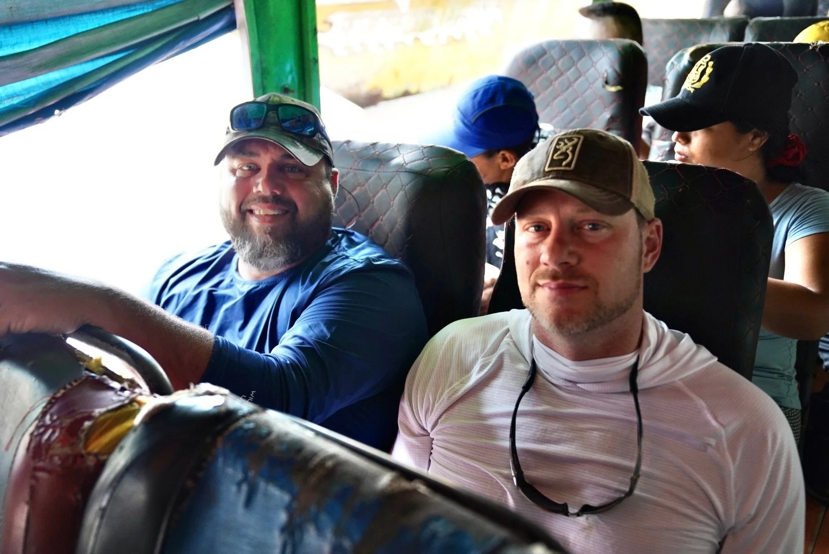 Two men sitting on a bus, smiling at the camera. One wears a baseball cap and with sunglasses on top, and the other has a cap and sunglasses hanging around his neck. There are other travelers sitting behind them.