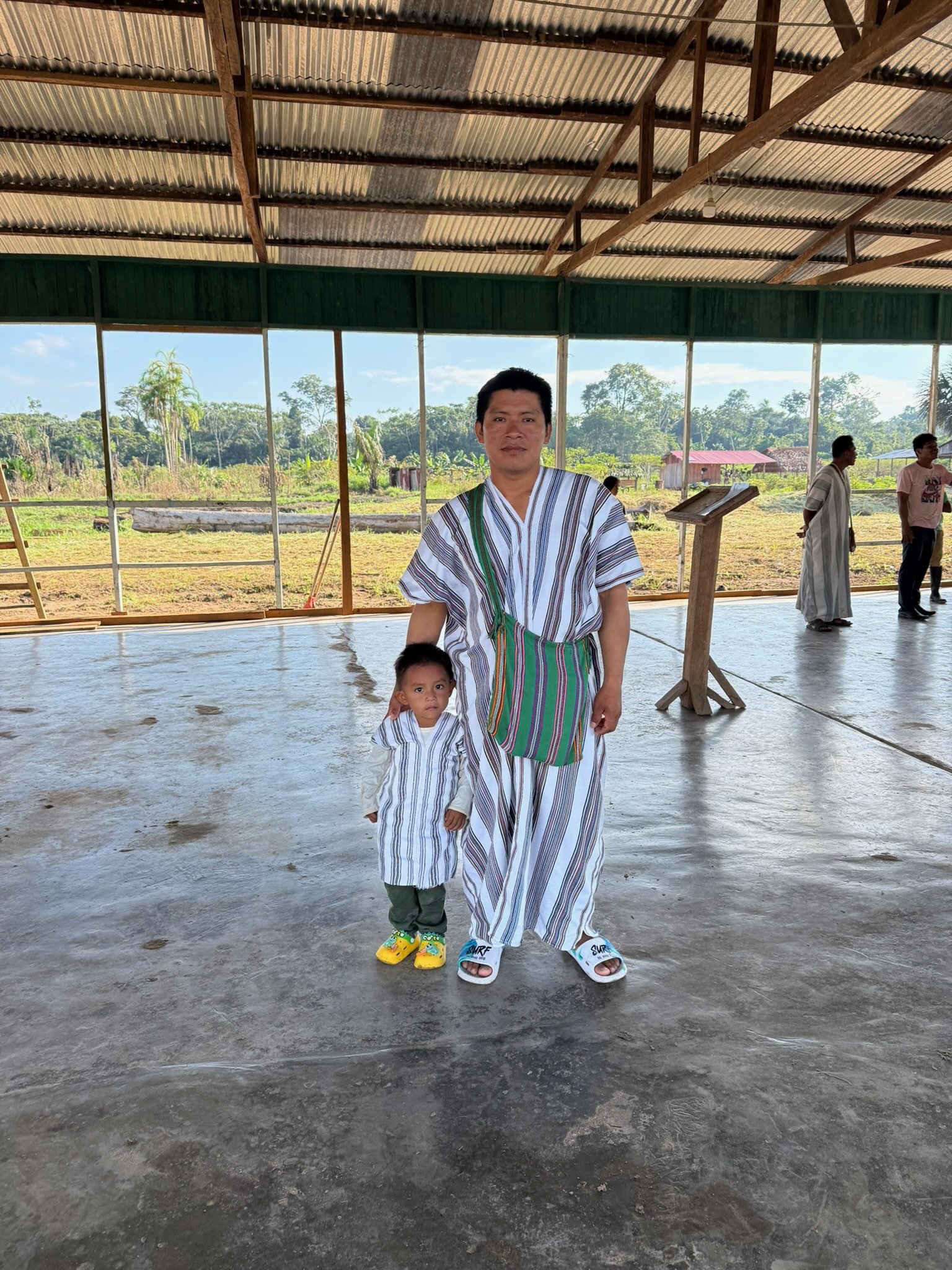 A man and a young boy dressed in matching traditional striped clothing standing inside a covered open-air hall, with a rural landscape visible through the large open windows behind them.