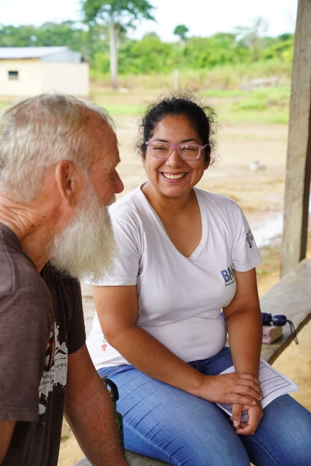 A smiling young woman with curly black hair, glasses, and a white t-shirt sitting next to an older man with white hair and beard, outdoors with a green landscape in the background.