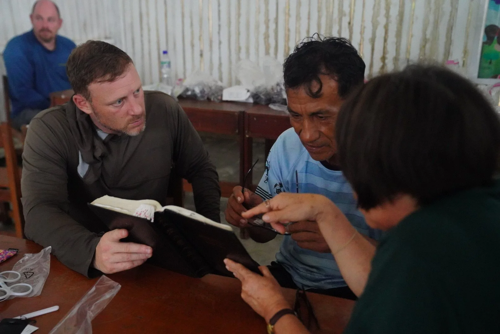 Four people sitting at a table talking, one showing something in a notebook, in a room with a corrugated metal wall.