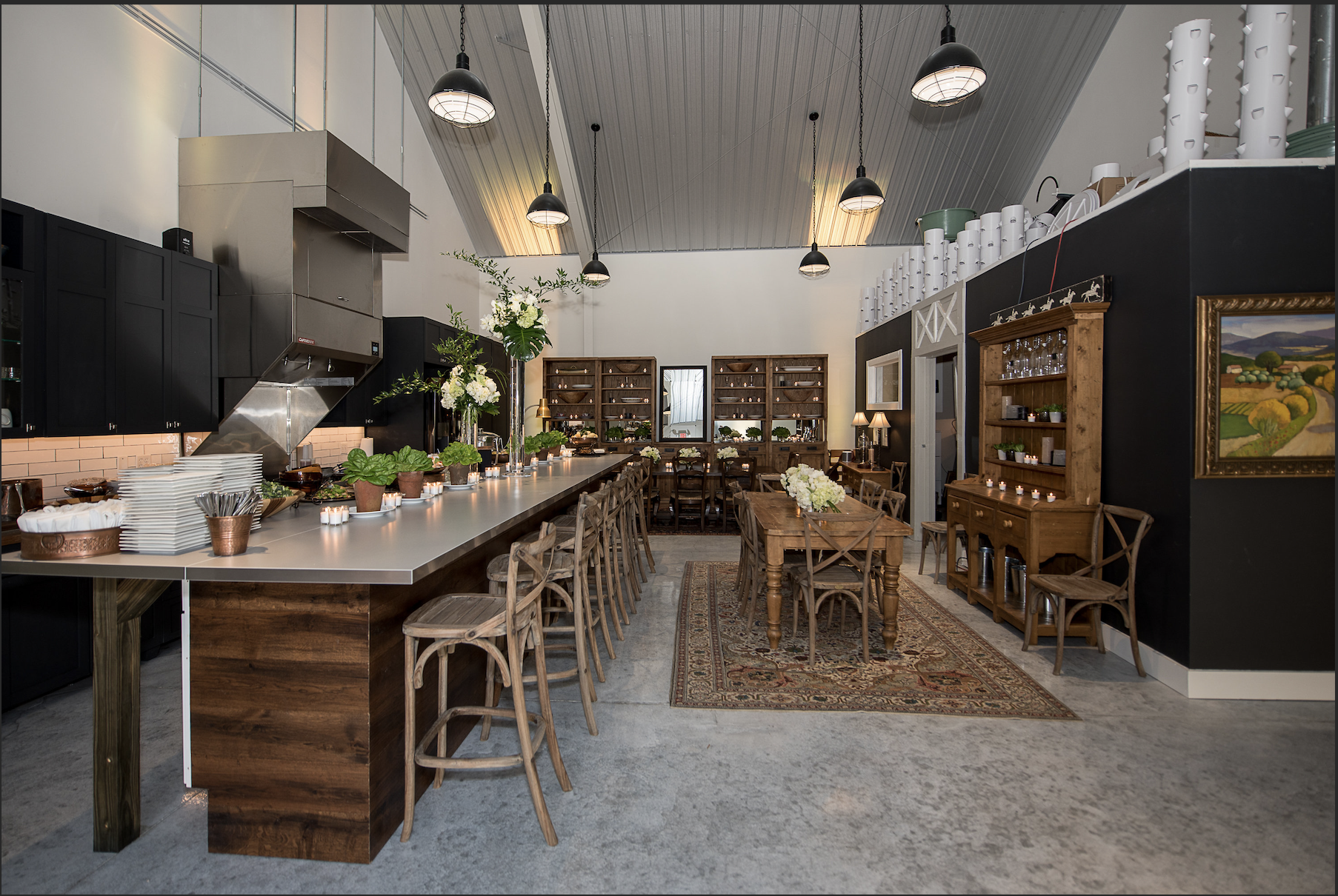 Interior of a cozy restaurant with a long kitchen counter and wooden tables, decorated with flowers and candles, featuring a mix of modern and rustic design elements.