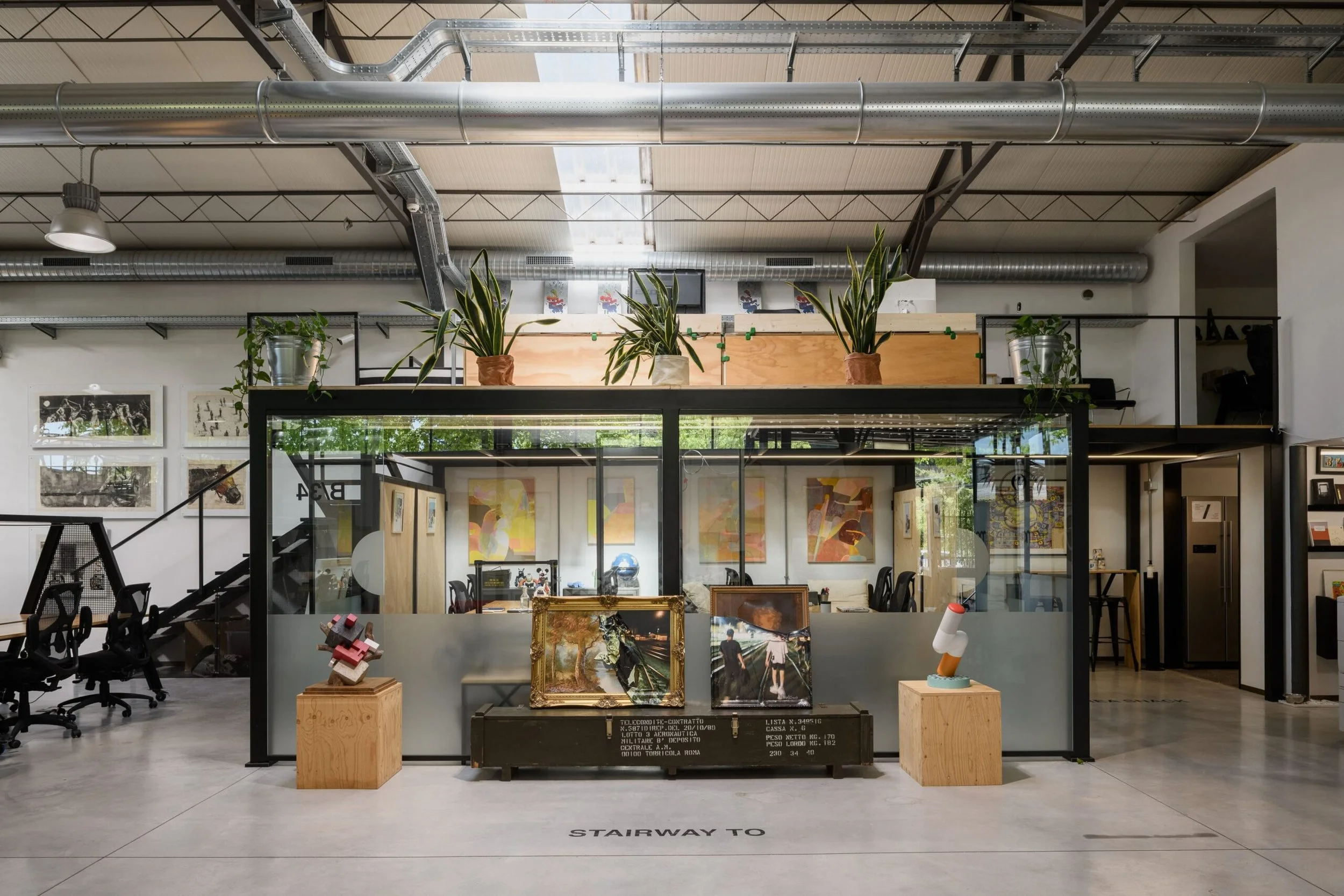 Interno dello spazio di coworking di Ala34 in Via di Affogalasino 34 a Roma. Con scrivanie, sedie e decorazioni artistiche, piante sullo sfondo e un'area con pavimento in cemento e scritte sul pavimento.