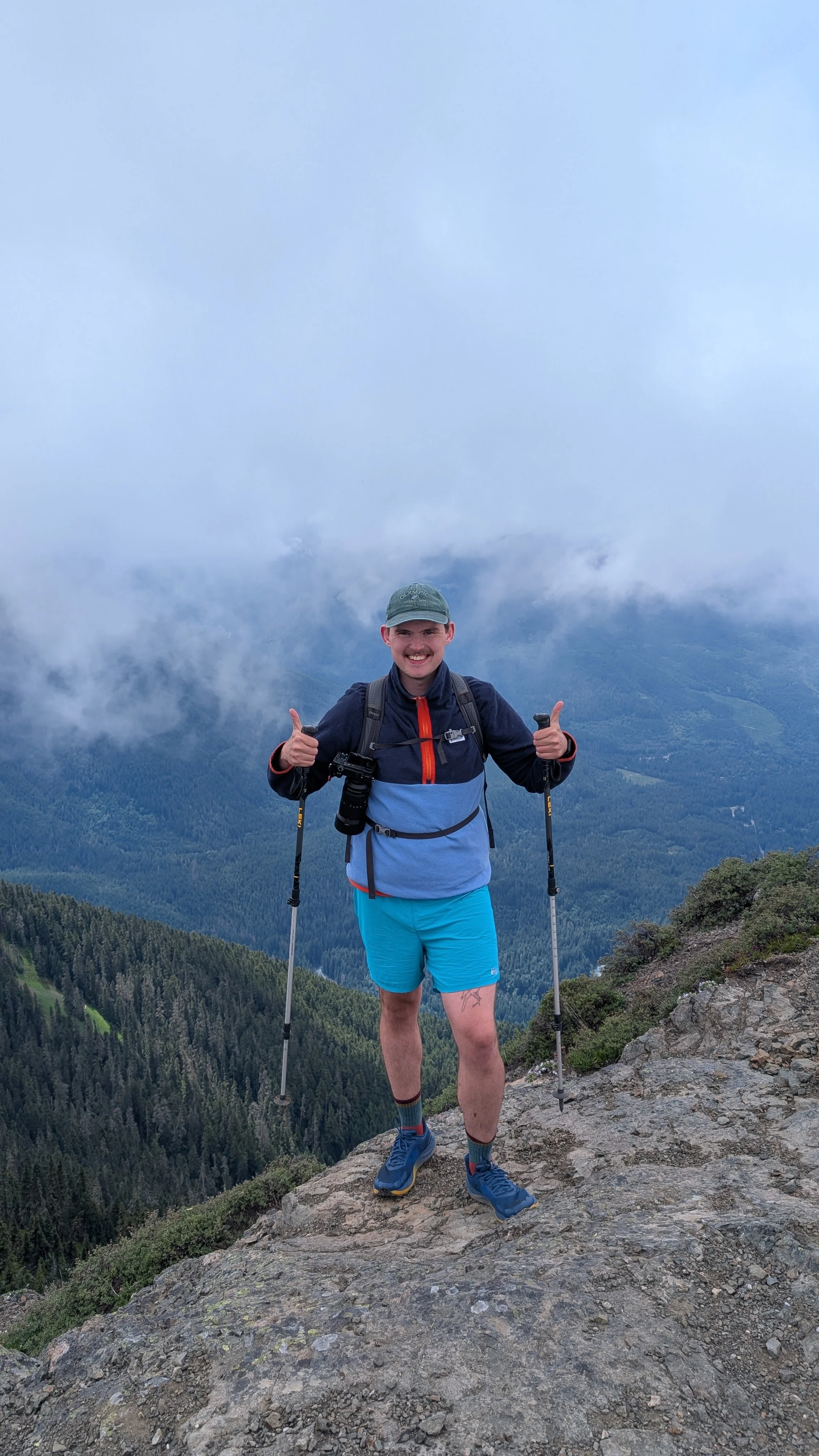 Young man standing on rocky mountain terrain with trekking poles, smiling at camera, with forested mountains and clouds in the background.