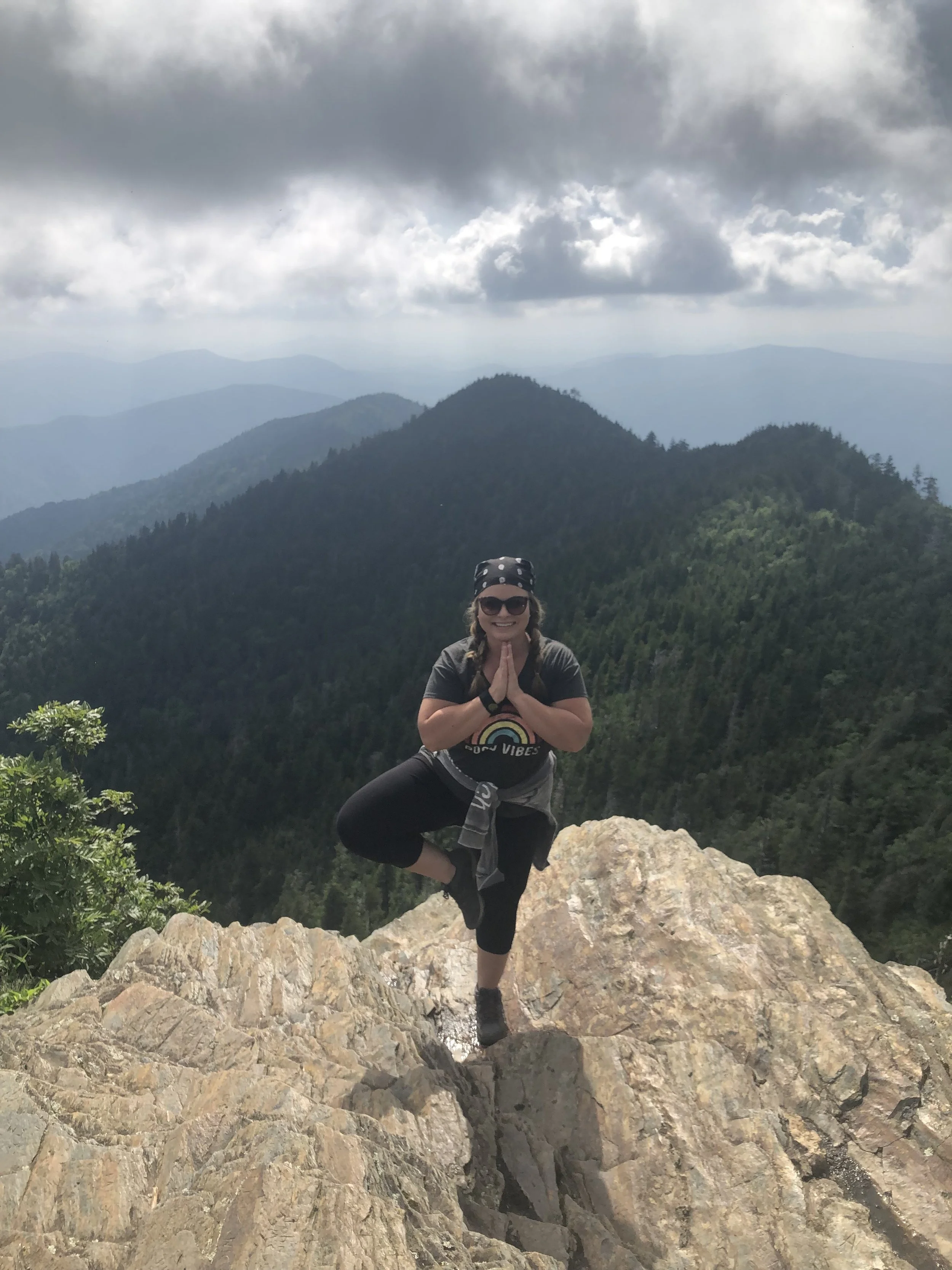 A woman practicing yoga in the tree pose on a rocky mountain peak with a forested mountain landscape and cloudy sky in the background.