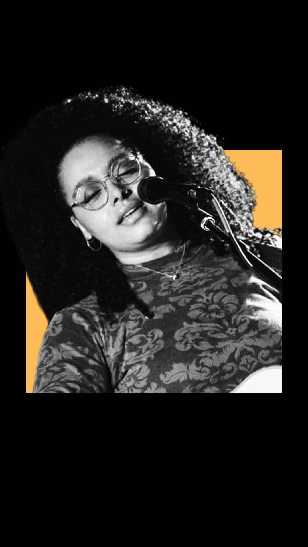 A young woman with curly hair and glasses singing or speaking into a microphone, wearing a patterned top, with a background that has black and yellow elements.