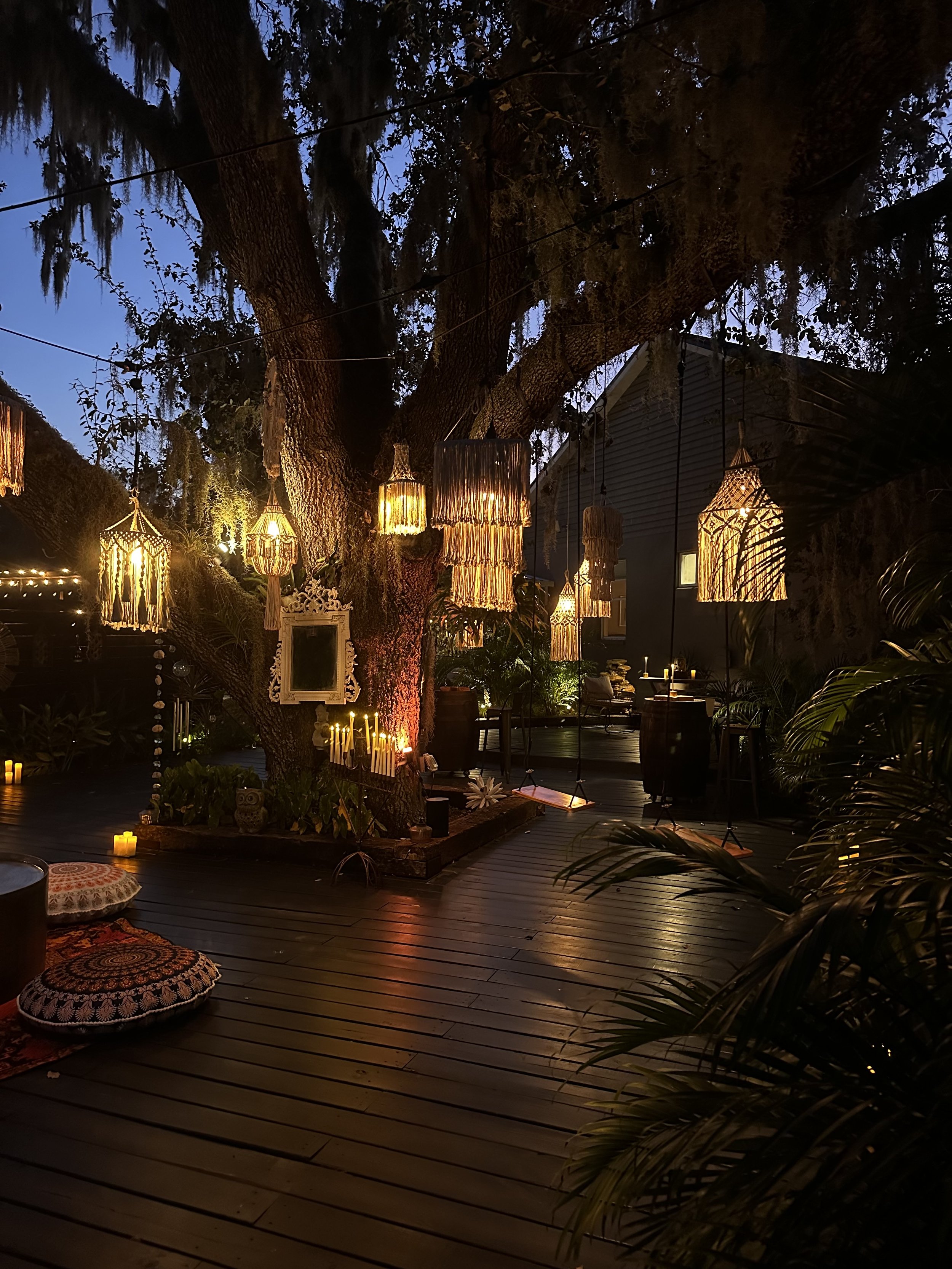Outdoor deck area decorated with hanging lanterns, candles, and cushions, with large tree in the center illuminated by warm lights at dusk.