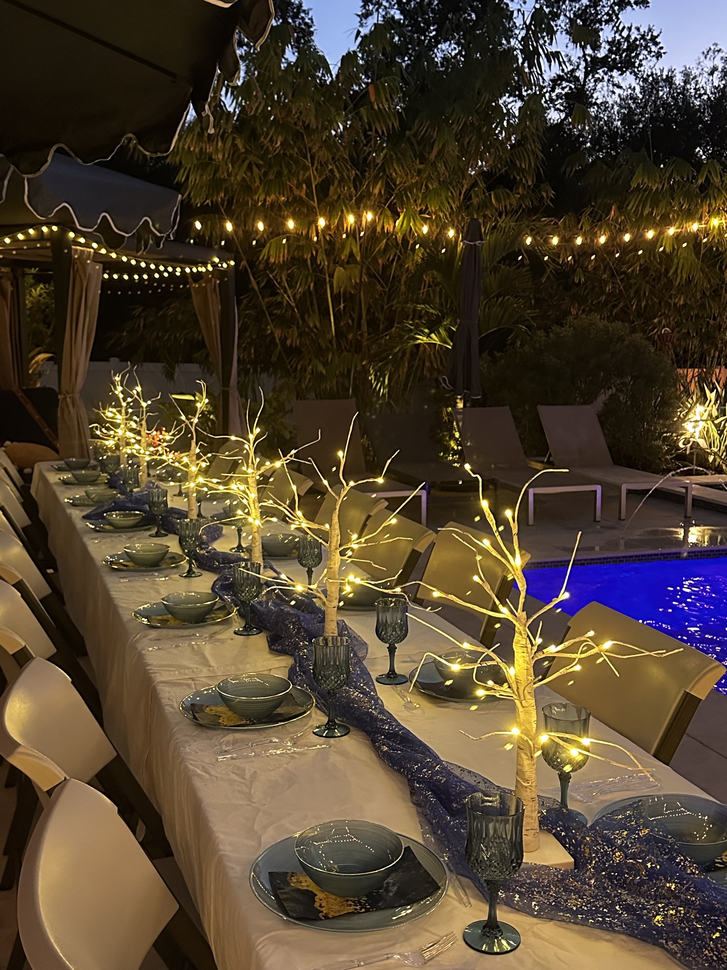 An outdoor evening dinner setup with a long table decorated with trees wrapped in fairy lights, set with bowls, plates, and glasses, near a pool with lounge chairs, surrounded by lush greenery and string lights overhead.